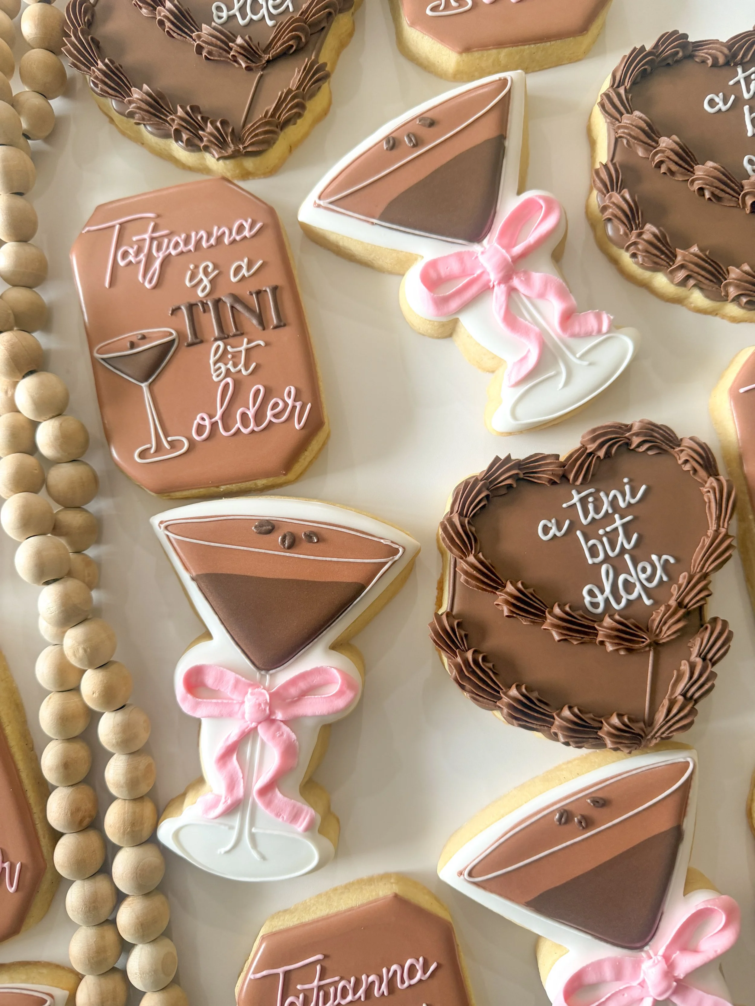 Decorative cookies with pink, brown, and white icing, some with text 'Tatyanna is a tiny bitolder' and others shaped like wine glasses with pink bows.