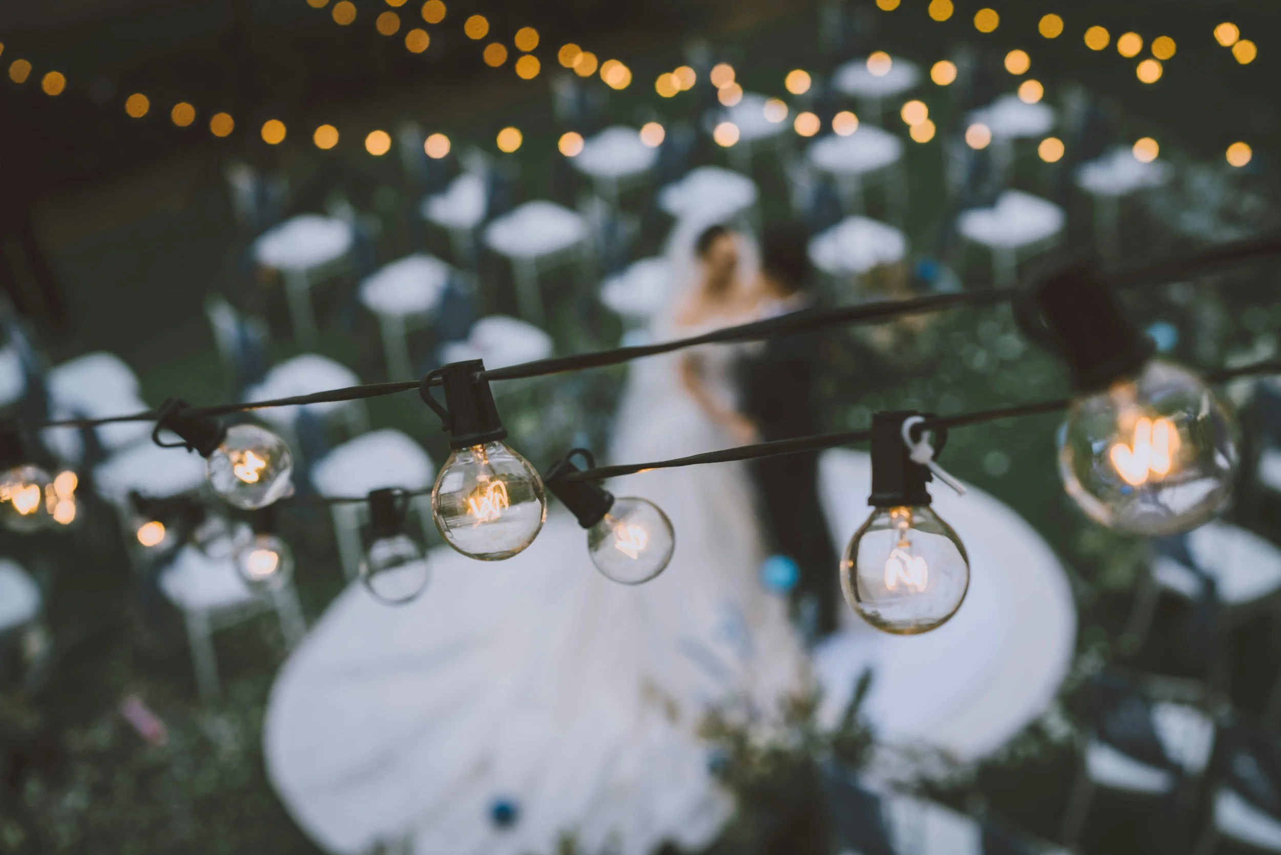 String of hanging string lights in the foreground with blurred people at a water fountain in the background.
