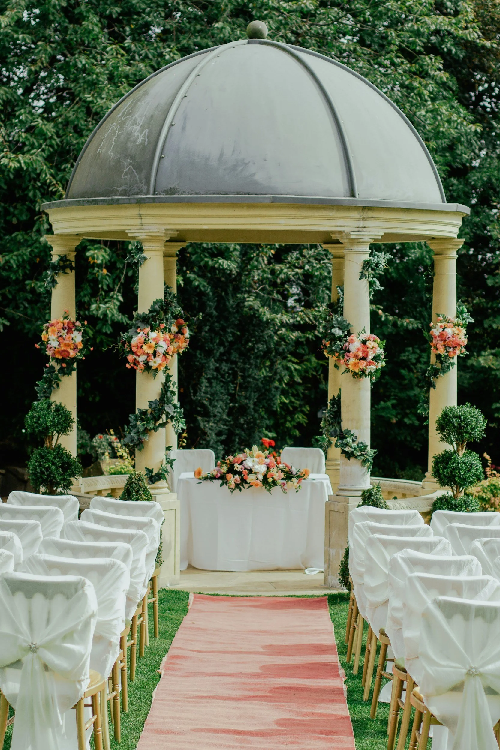 Outdoor wedding ceremony setup with white chairs, floral decorations, a pink aisle runner, and a covered pavilion with columns and a floral table centerpiece.
