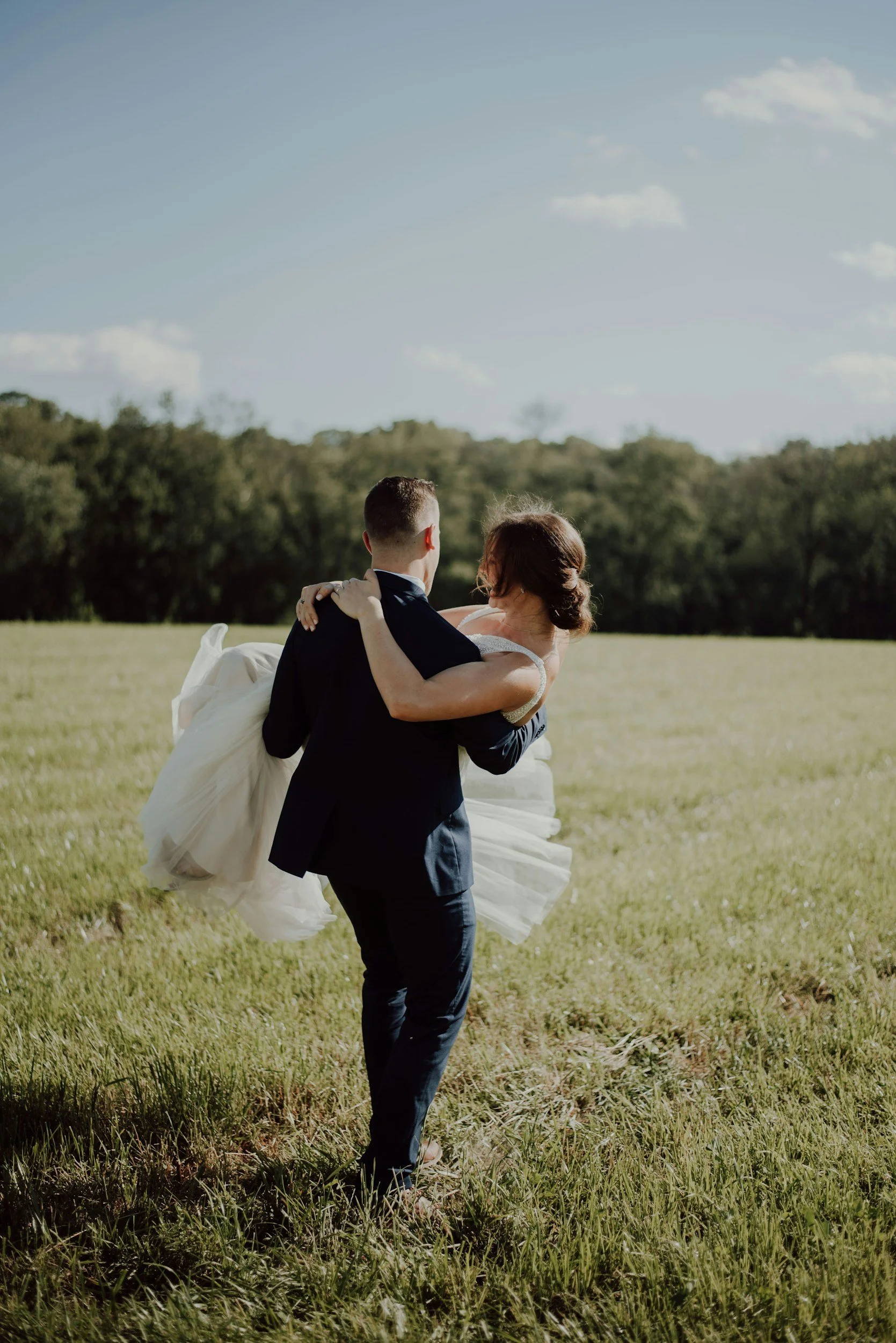 A groom in a black suit carrying a bride in a white wedding dress across a grassy field with trees in the background under a partly cloudy sky.