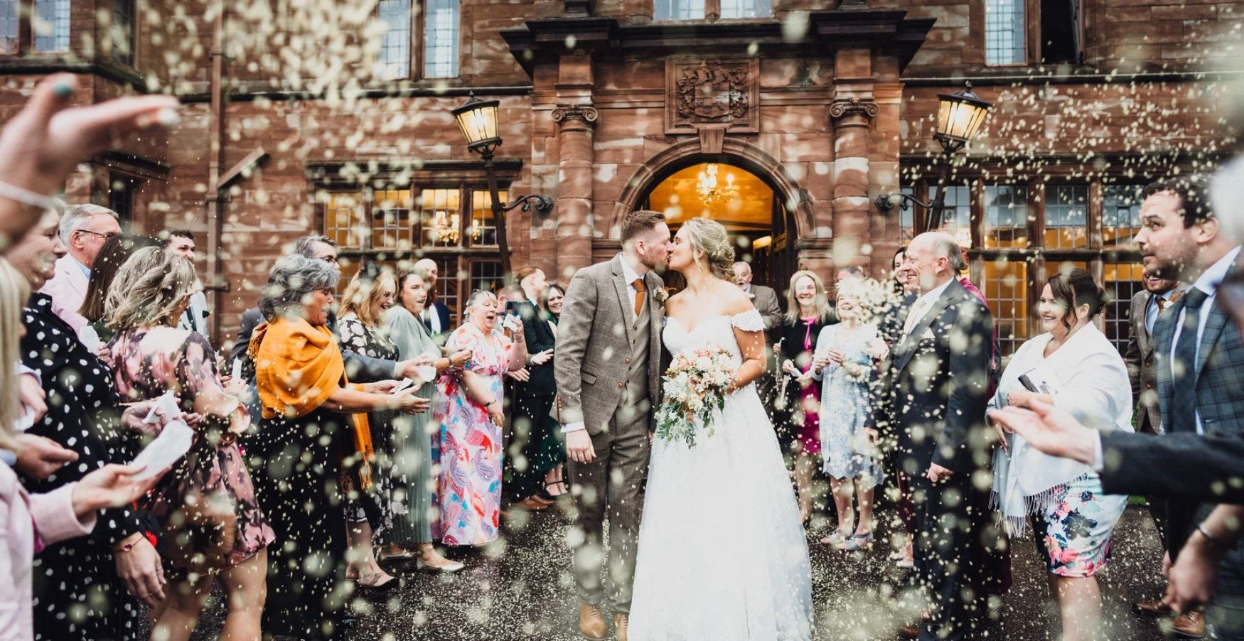 A newlywed couple kisses outside a brick building during their wedding celebration, with guests throwing confetti around them.