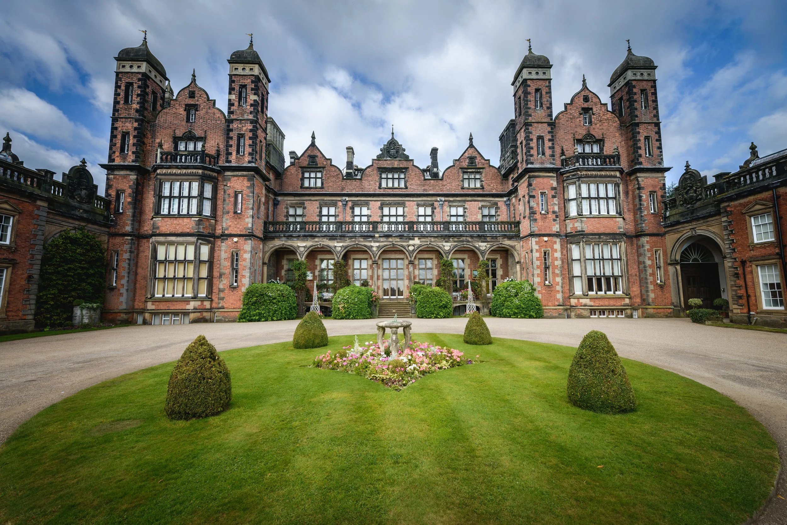 Front view of a large historic red brick castle with multiple turrets, arched windows, and a courtyard garden with green bushes, a fountain, and flowering plants, under a daytime cloudy sky.