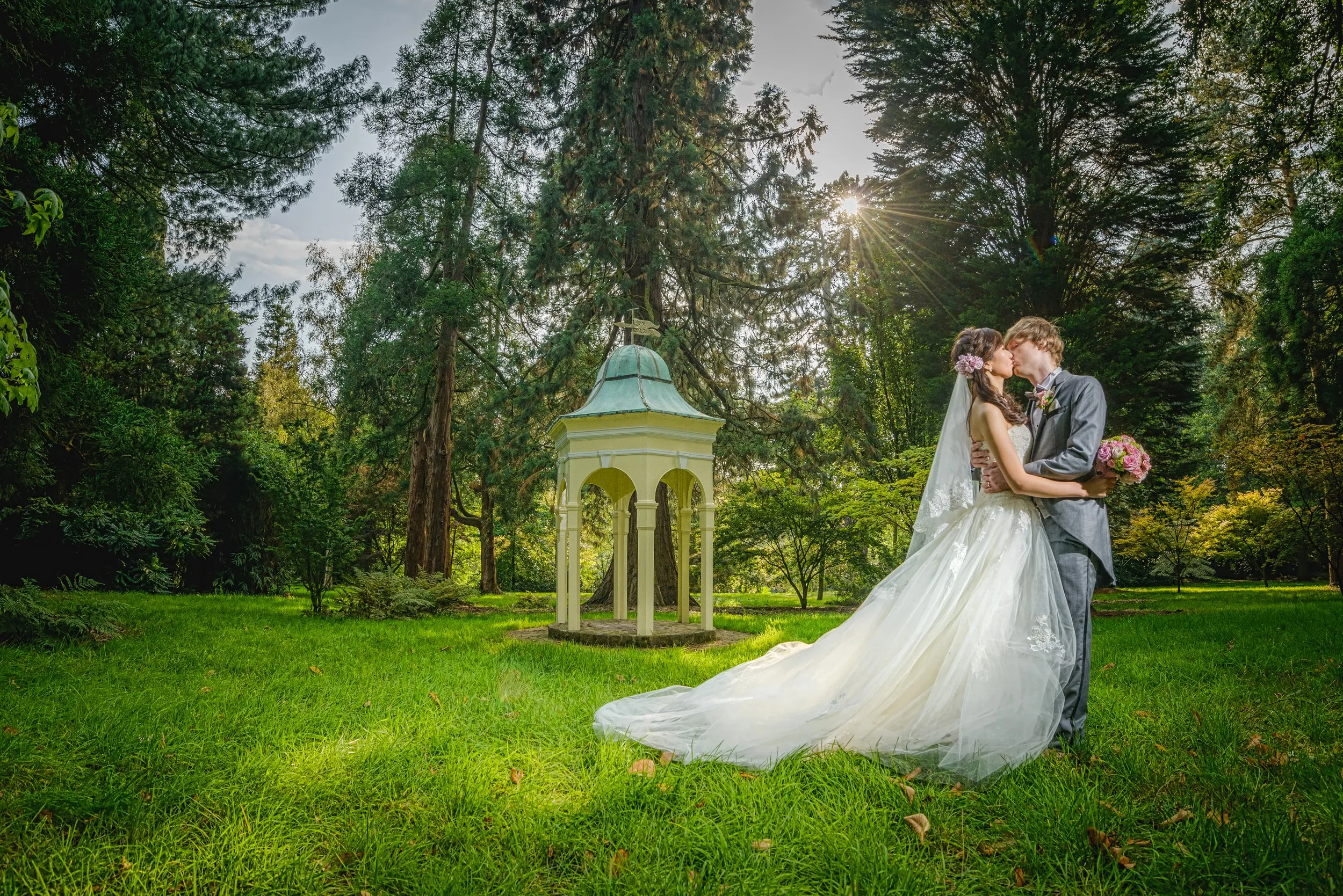 A bride and groom sharing a kiss outdoors in a green park, surrounded by tall trees, with the sun shining through the branches and a small white pavilion in the background.