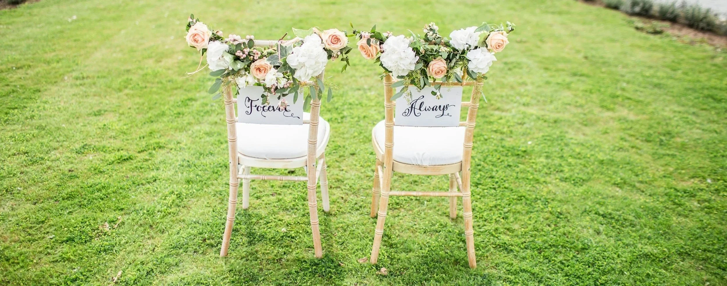 Two chairs decorated with floral arrangements, each with a sign that reads 'Forever' and 'Always,' set on a grassy outdoor area.