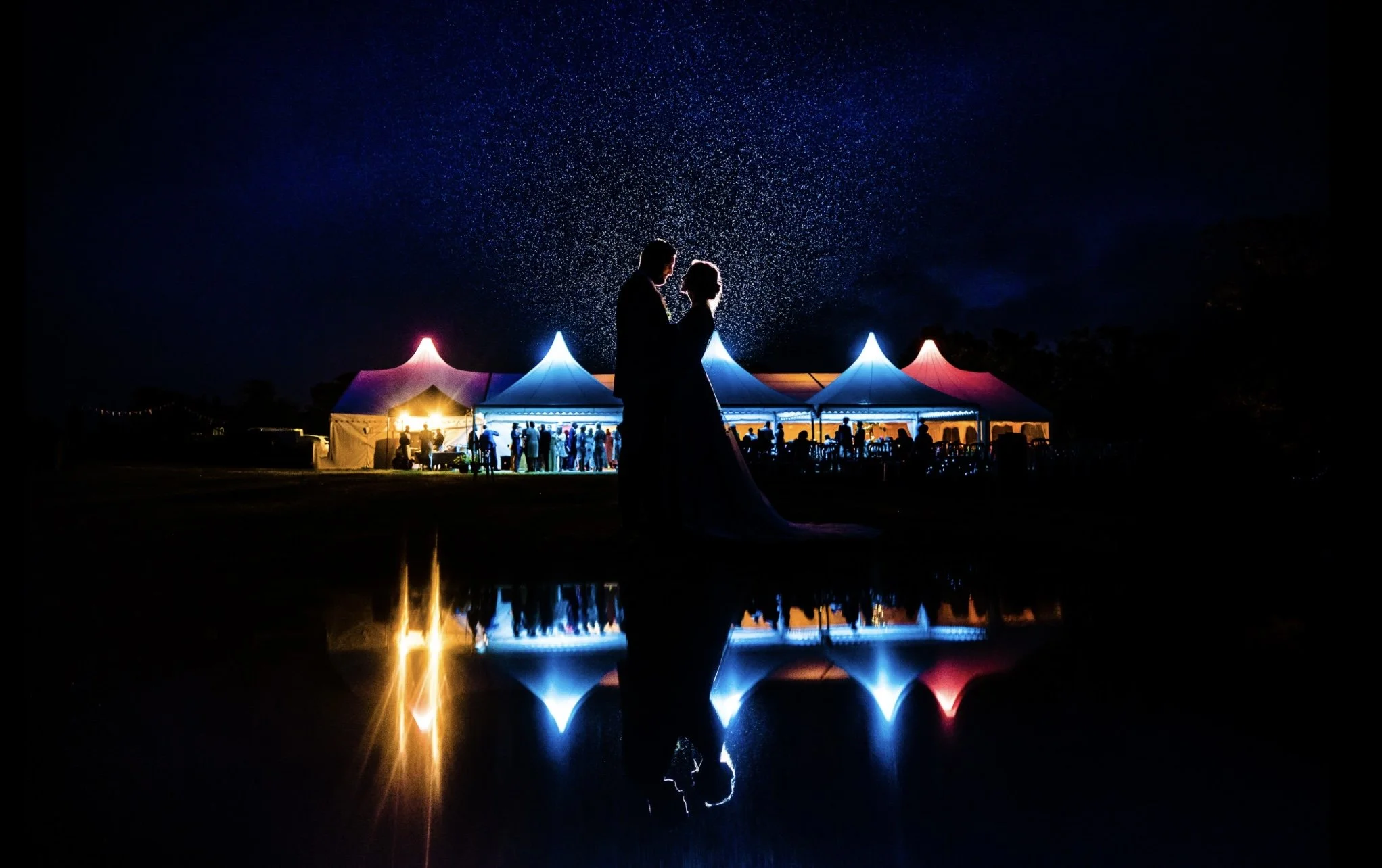 Silhouette of a couple dancing under a starry sky at night, with colorful illuminated tents nearby, their reflection visible in a body of water.