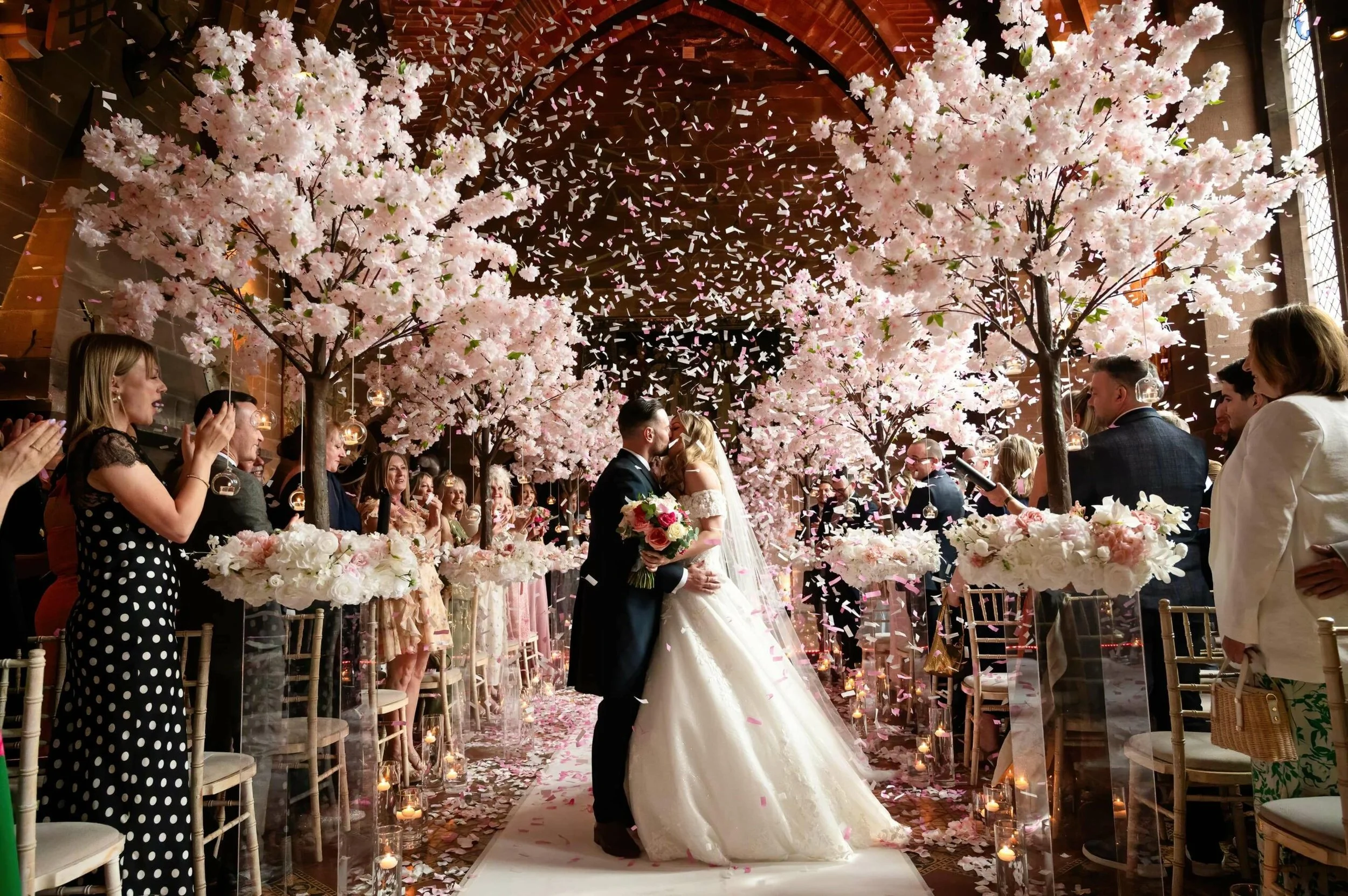 A wedding ceremony with the bride and groom kissing under pink cherry blossom trees, confetti falling, and guests applauding and smiling in a decorated indoor space.