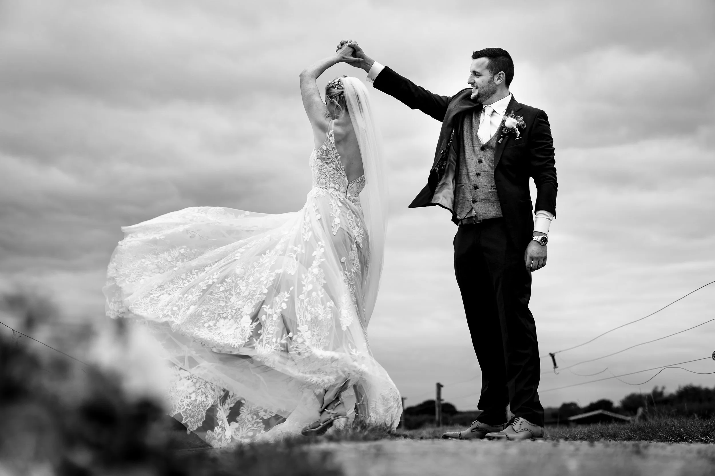 Black and white photo of a bride and groom dancing outdoors. The bride is wearing a lace wedding dress with a veil and the groom is dressed in a tuxedo with a boutonniere, holding the bride's hand.
