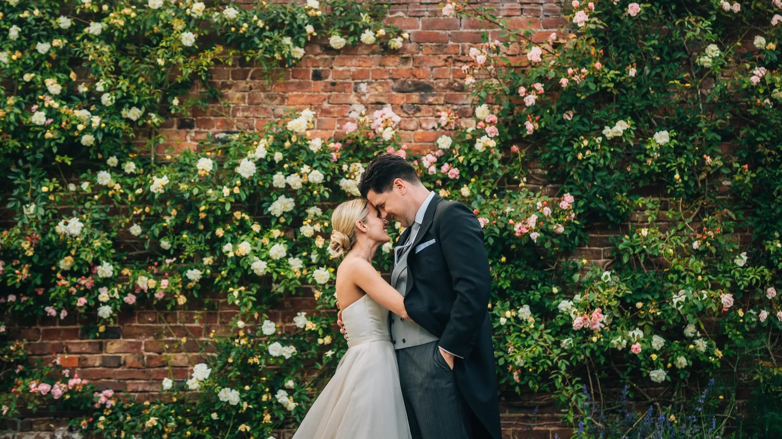 A couple dressed in wedding attire embracing and touching foreheads in front of a brick wall covered with white and pink flowers.