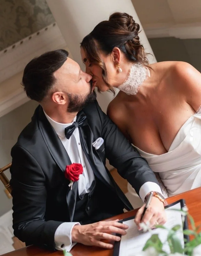 A newlywed couple sharing a kiss, with the groom in a black tuxedo with a red rose boutonniere, and the bride in a white wedding dress and lace choker.