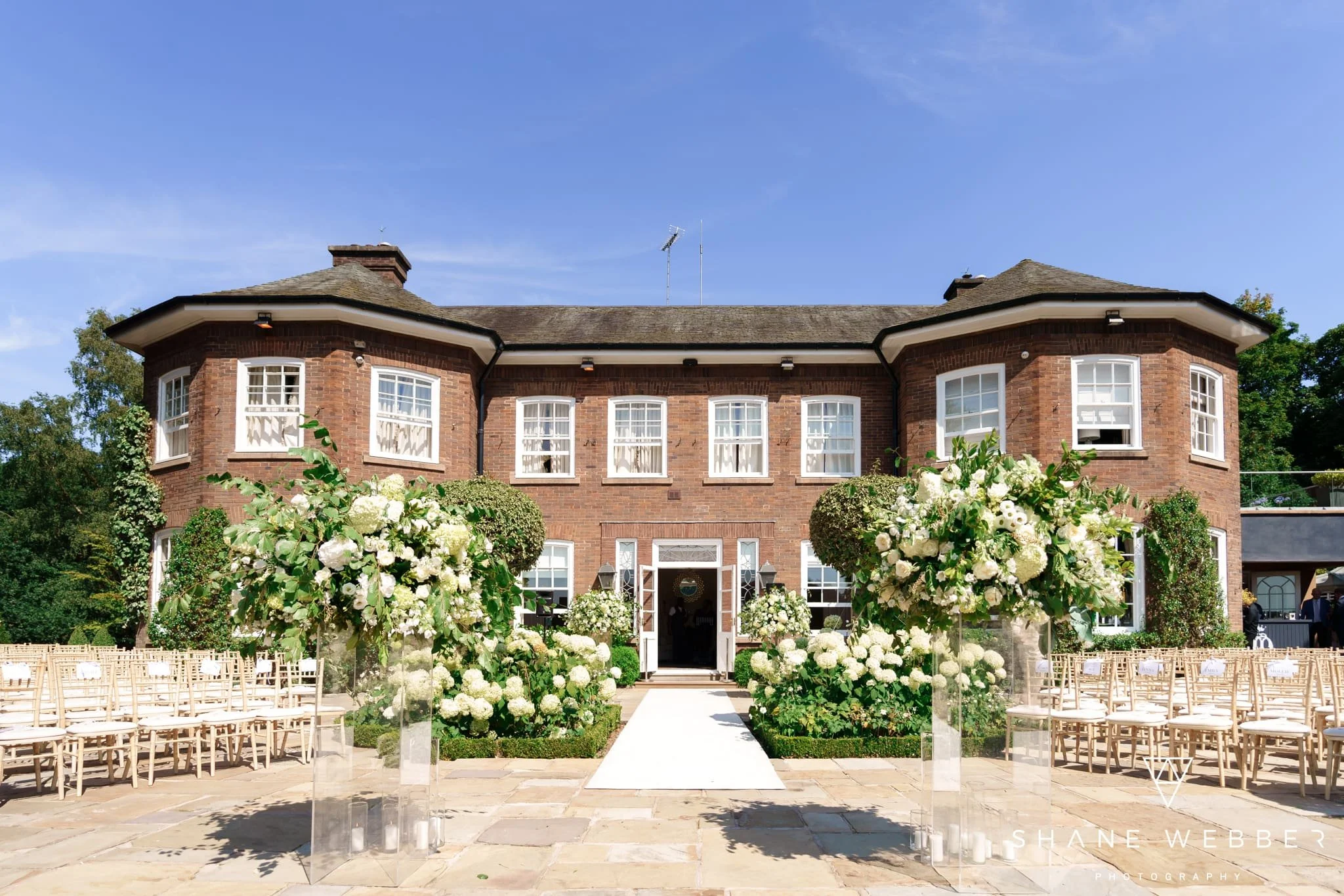 A brick house decorated for a wedding ceremony with chairs arranged on the lawn, white flower arrangements, and a white aisle runner leading to the entrance.