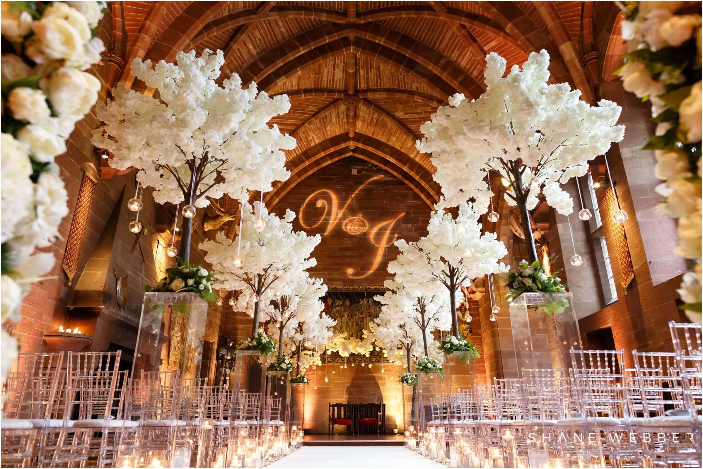 Indoor wedding ceremony setup with white flowering trees and hanging lights, featuring clear chairs arranged along a white aisle, in a venue with brick arches and warm lighting, and a monogrammed sign at the end of the aisle.