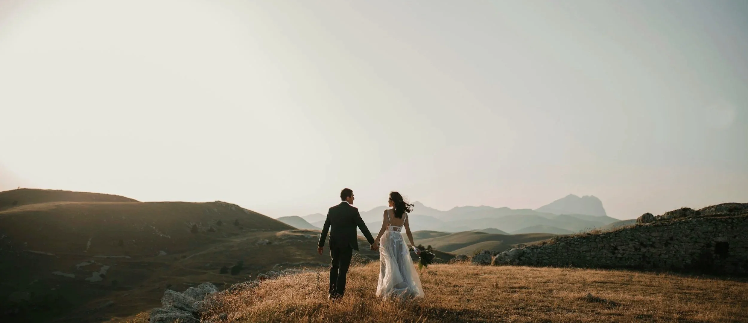 A couple dressed in wedding attire holding hands and walking in a scenic valley at sunset, with mountains in the background.