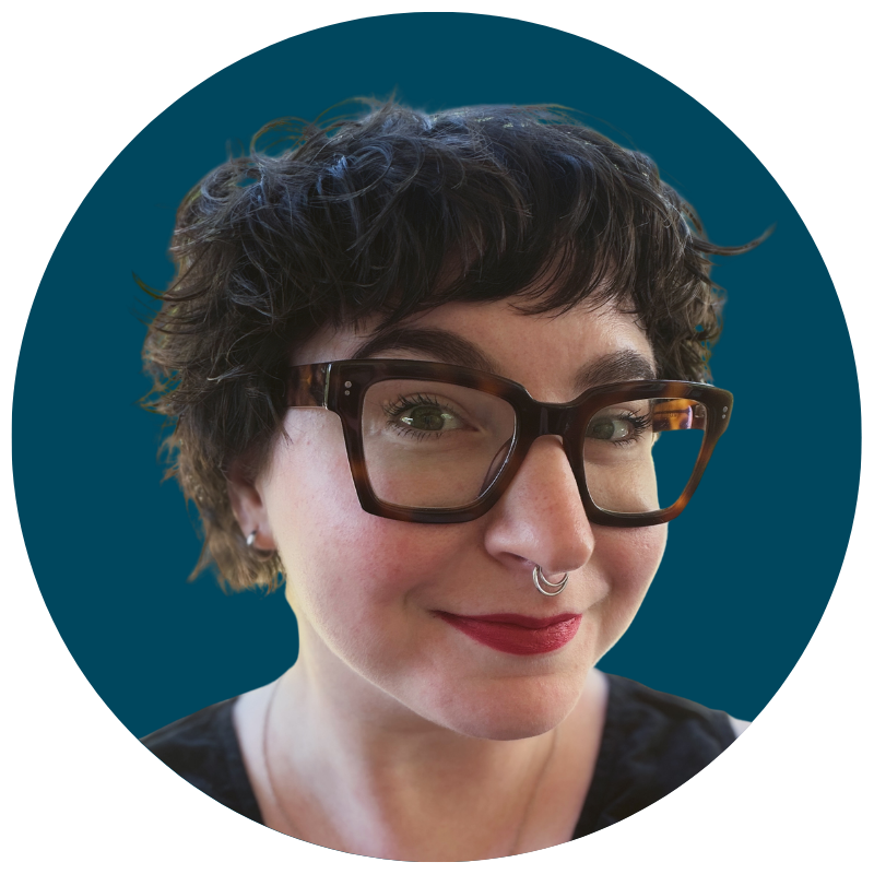 A woman with short curly dark hair, wearing large dark glasses, a nose ring, red lipstick, and a black top, smiling at the camera against a teal background.