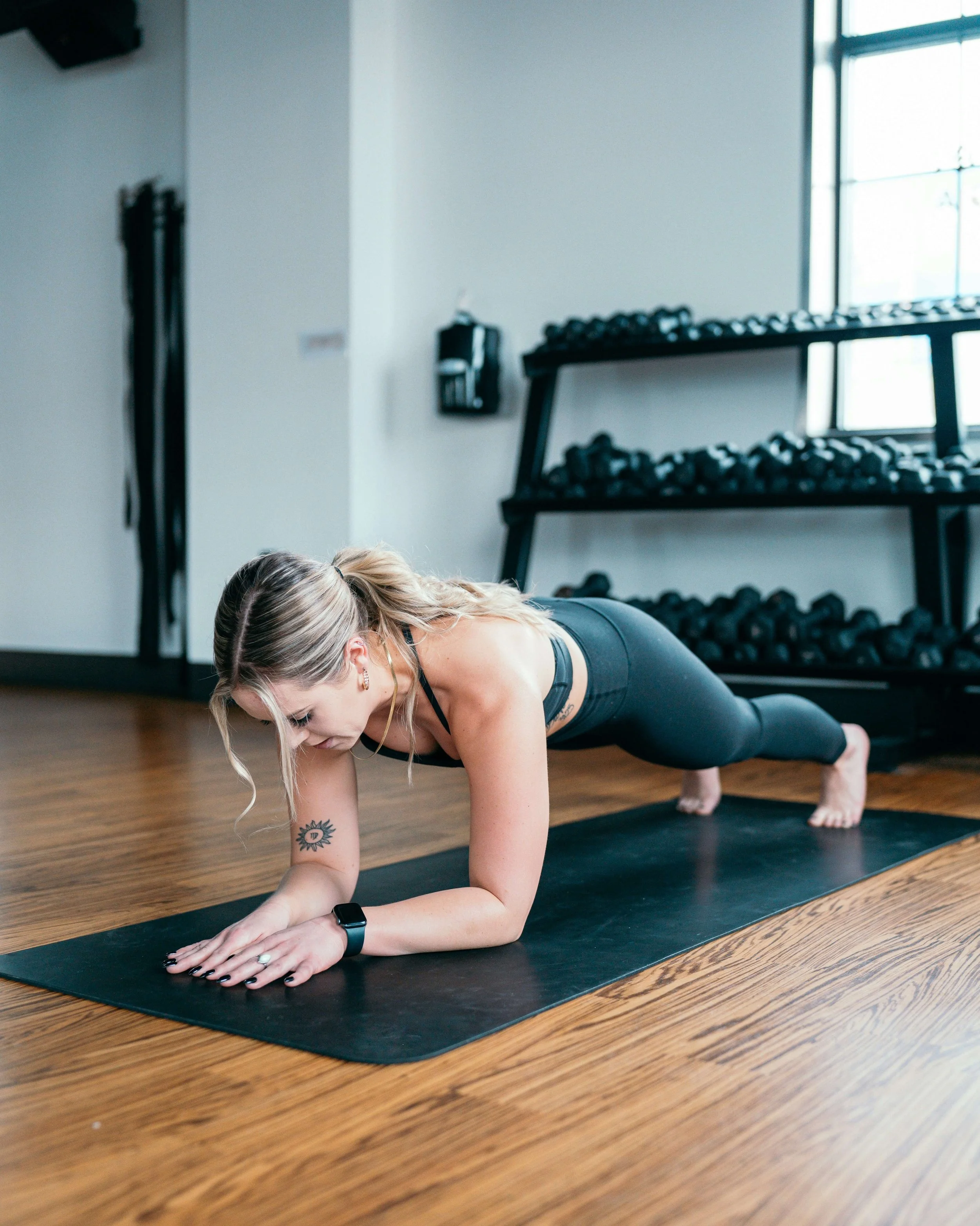 A woman with blonde hair in a ponytail and tattoos on her arm is doing a plank exercise on a black yoga mat in a gym, wearing black workout clothes and a smartwatch, with stacked dumbbells and windows in the background.