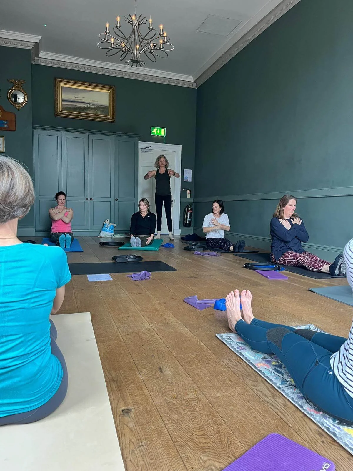 A group of women participating in a yoga class, sitting on mats with some stretching or relaxing poses, led by an instructor who is standing and demonstrating.