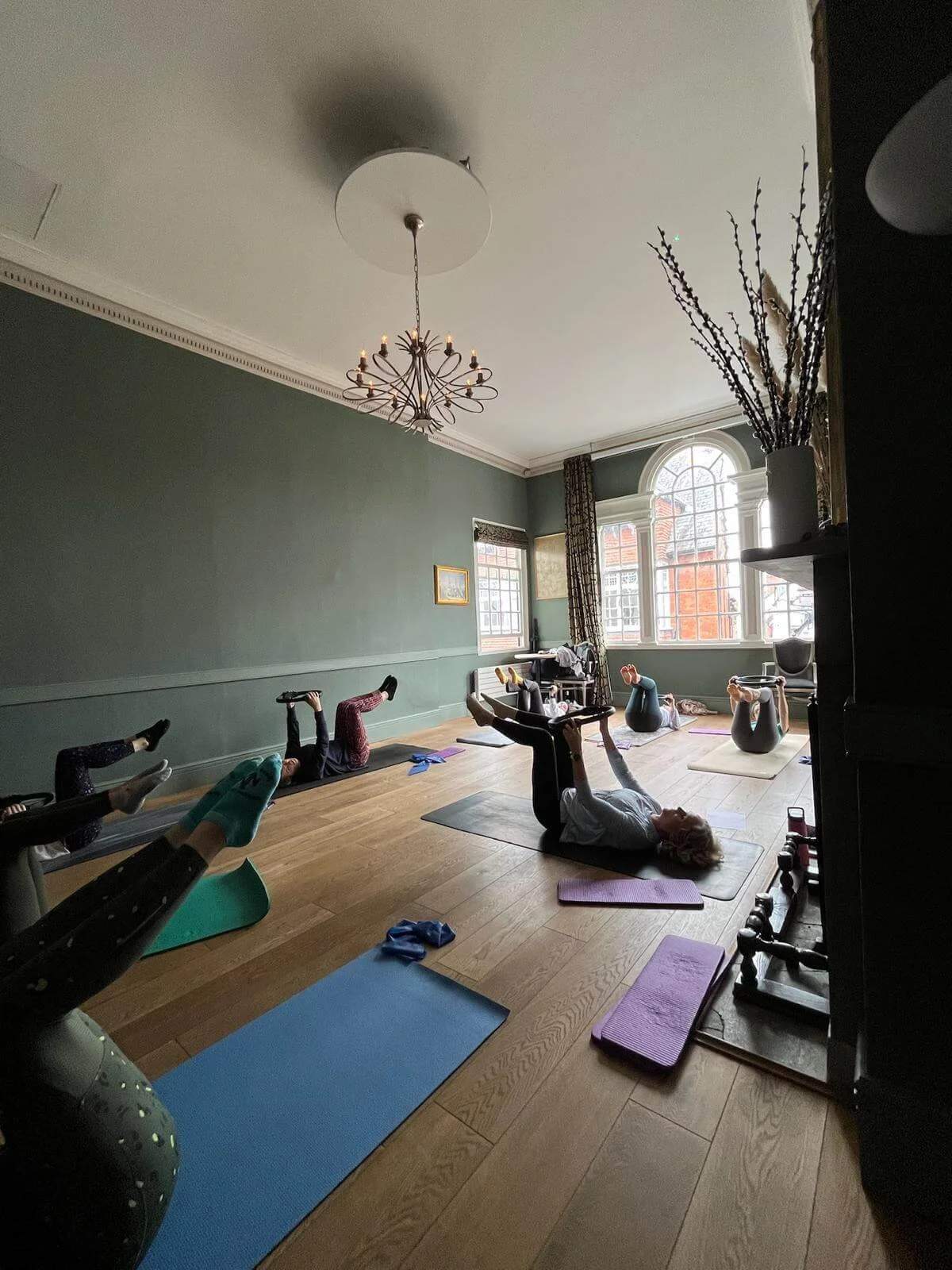 Group of people participating in a pilates class in a spacious, well-lit room with large windows, wooden flooring, and a chandelier hanging from the ceiling. They are lying on pilates mats, holding their legs in the air.