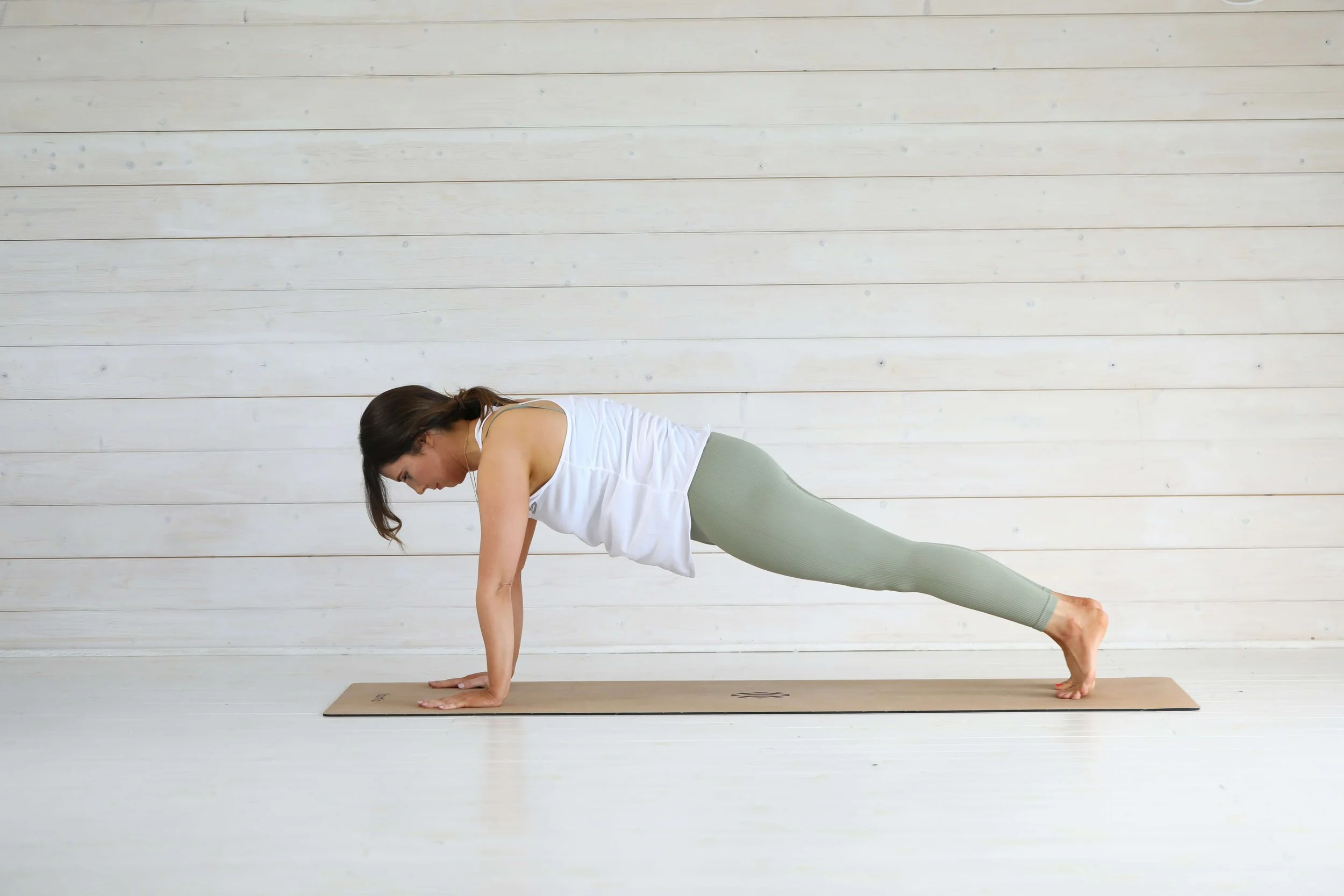 A woman practicing yoga indoors, performing a plank pose on a yoga mat against a white wooden wall.