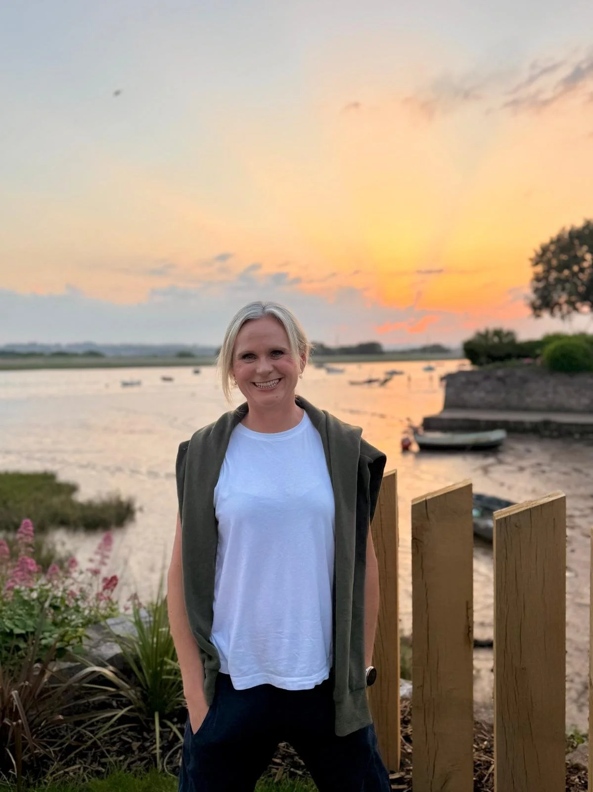 Beth of Exe Valley Pilates smiling outdoors during sunset near water with boats, trees, and a wooden fence in the background.