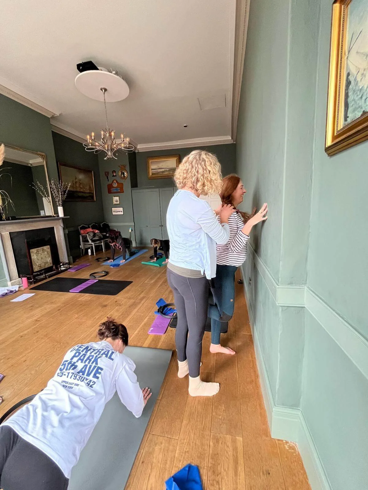 Two women standing and practicing pilates against a green wall while children do yoga on mats in a living room with wooden flooring, a fireplace, and artwork on the walls at Exe Valley Pilates