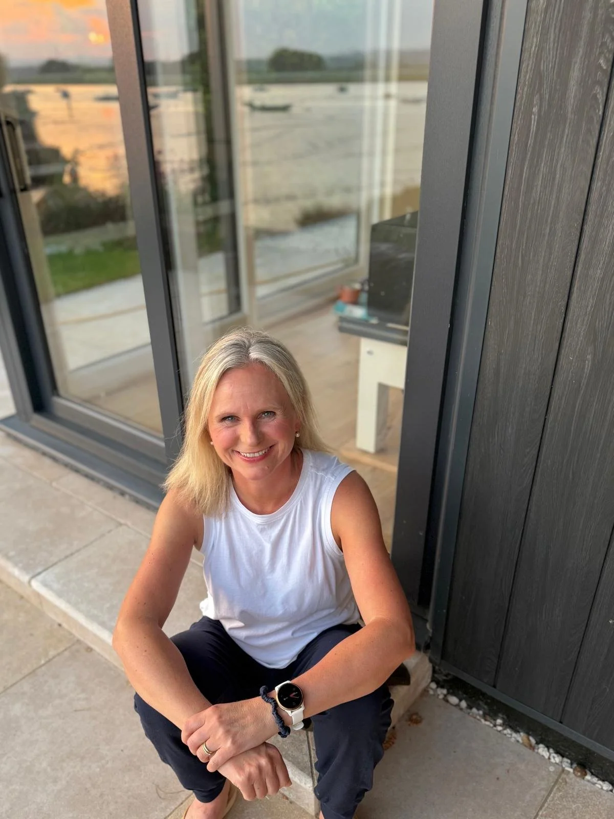 Beth of Exe Valley Pilates with blonde hair, wearing a white sleeveless top and black pants, sitting outdoors on a concrete step near large glass sliding doors, with a view of a river and boats in the background during sunset.