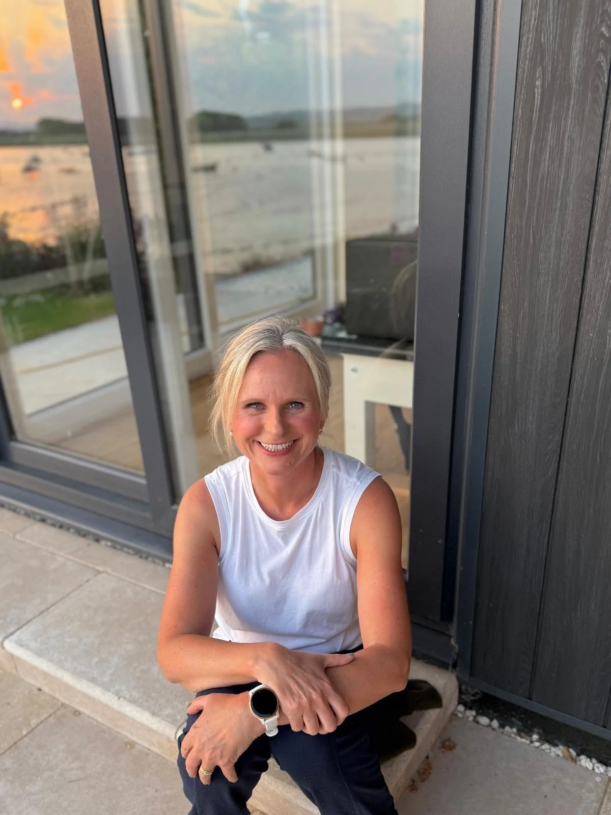 Beth of Exe Valley Pilates with short blonde hair, wearing a white sleeveless top, sitting on a stone step outside near a black-paneled building with large glass windows, during sunset water views in the background.