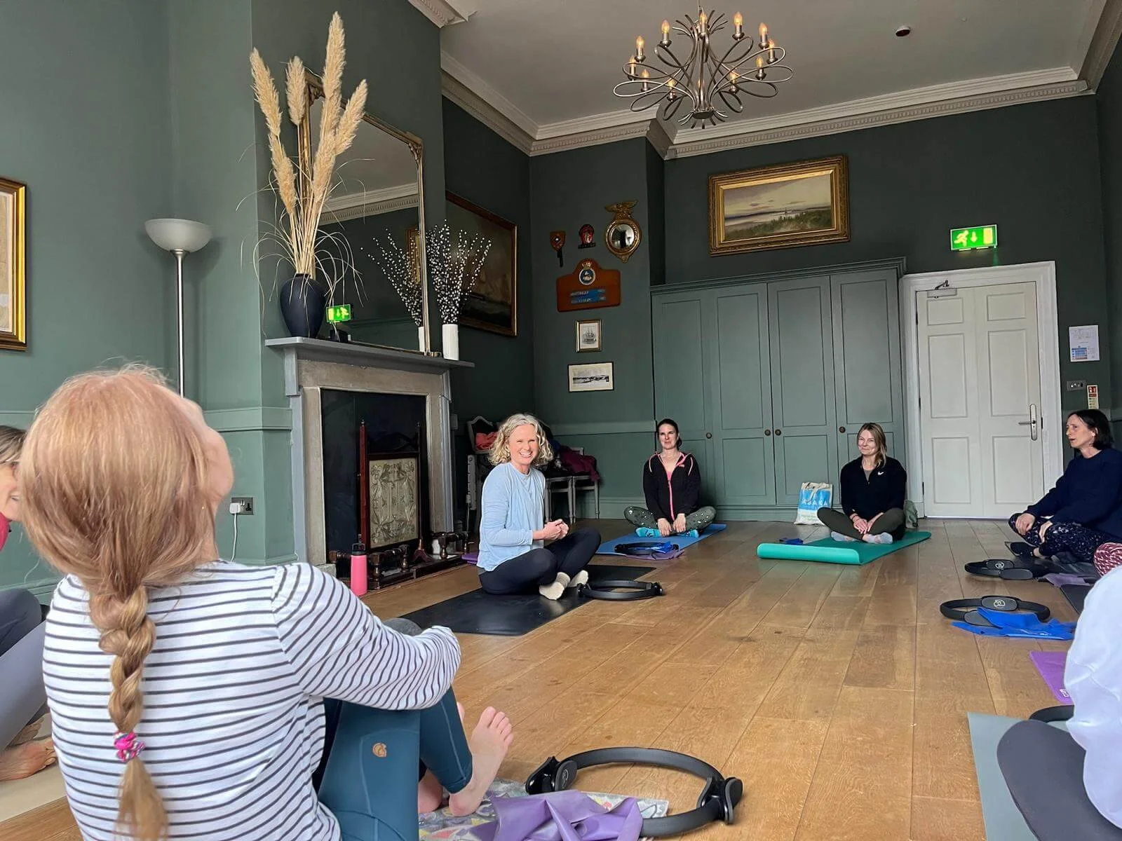 Women participating in a pilates class in a spacious room with green walls, a fireplace, and artwork, sitting on mats and listening to Beth of Exe Valley Pilates