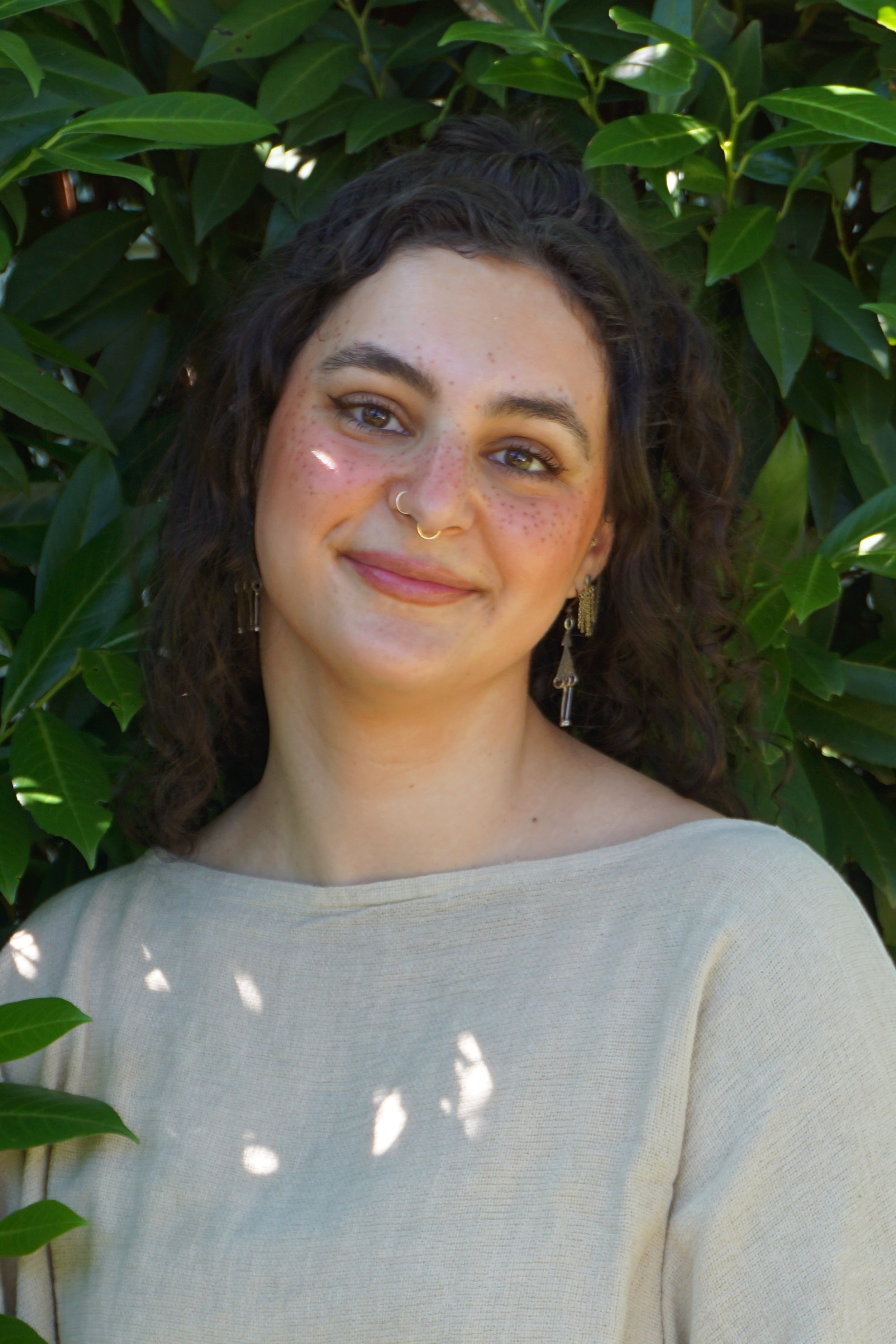 A young woman with curly dark hair, freckles, and tan skin, smiling while standing in front of green leafy bushes, wearing a beige top and gold earrings.