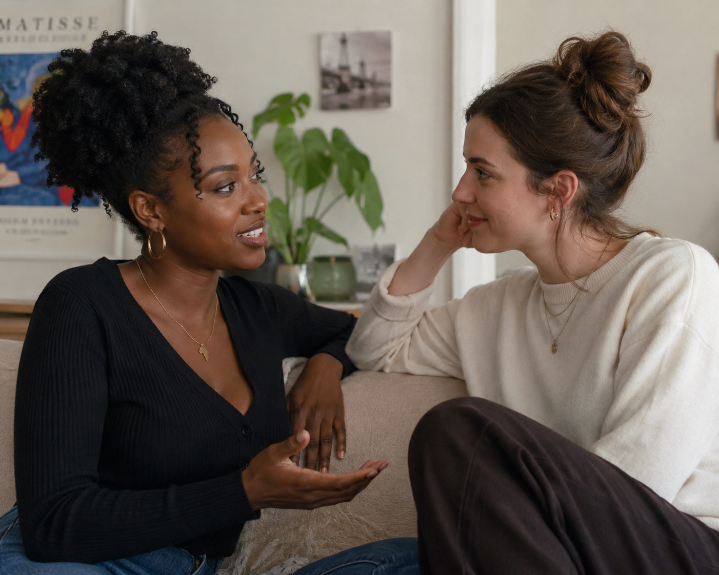 Two women engaging in conversation sitting on a beige couch in a cozy room, with artwork and plants in the background.