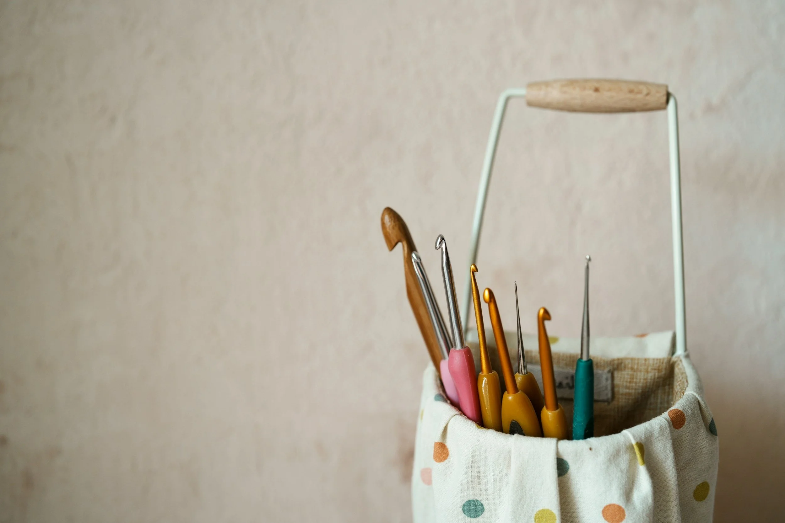 A fabric storage bin with multicolored polka dots holding several knitting or crochet hooks of various sizes and colors, with some hooks having wooden or metal handles, against a plain wall background.
