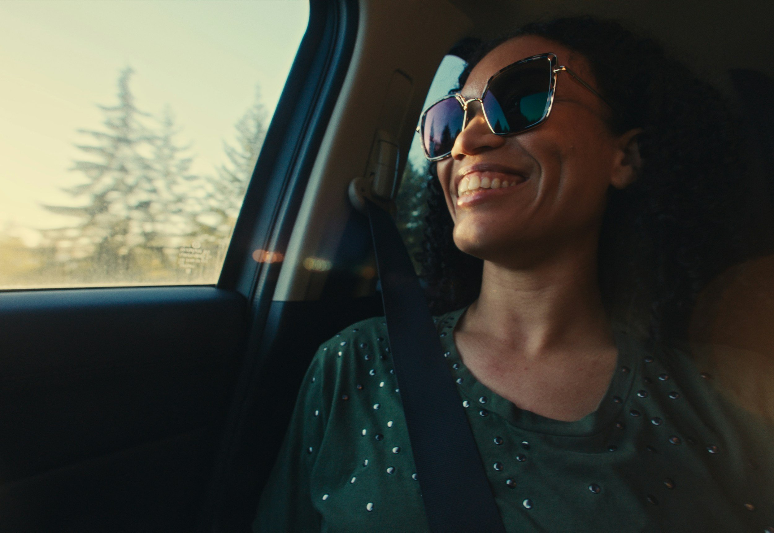 Mujer con gafas de sol coronando una sonrisa en un vehículo, vista desde el interior con árboles borrosos por la ventana.