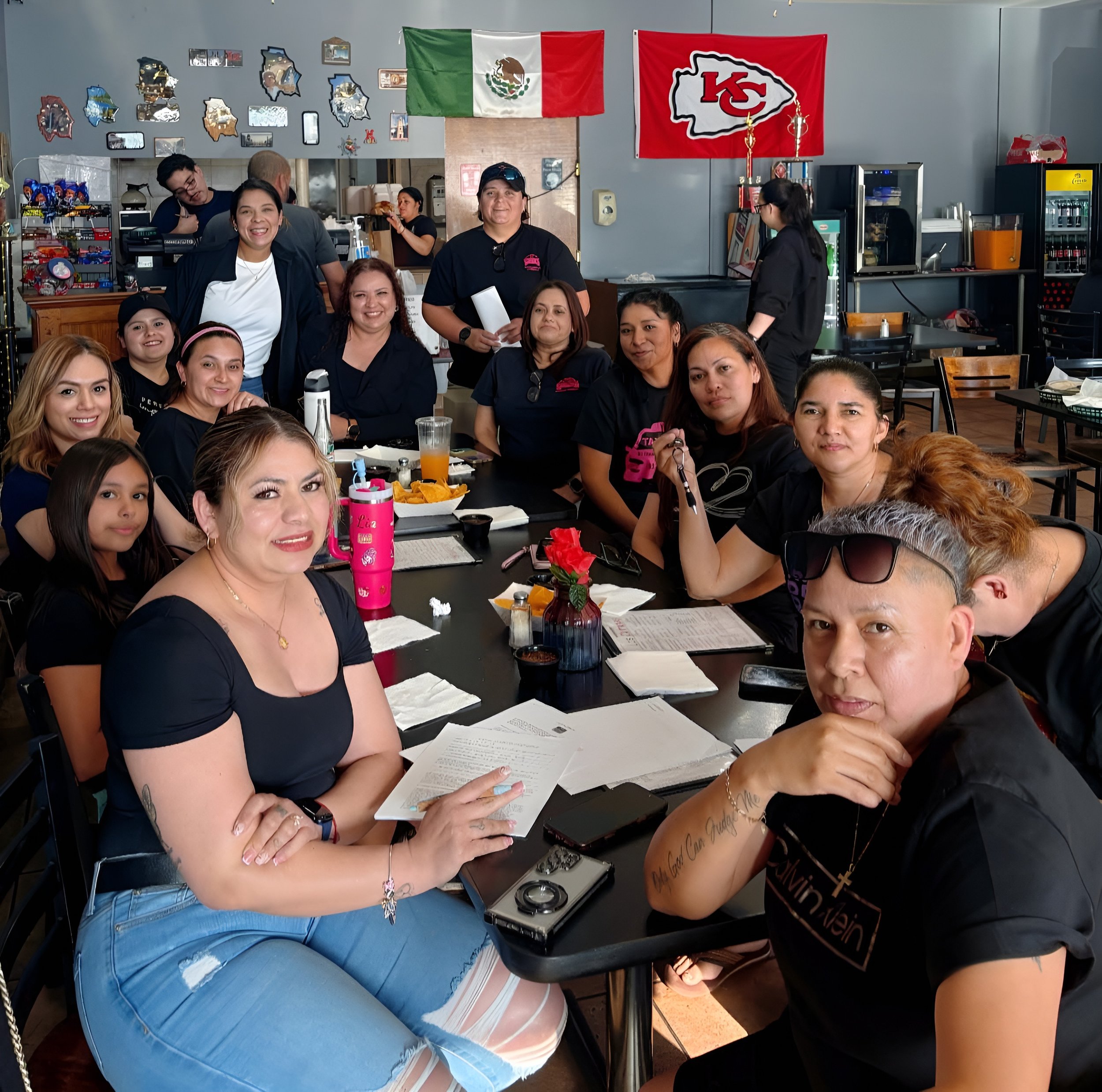 Group of diverse smiling people gathered around a table with food and drinks inside a restaurant decorated with Mexican flags and pictures on the wall.