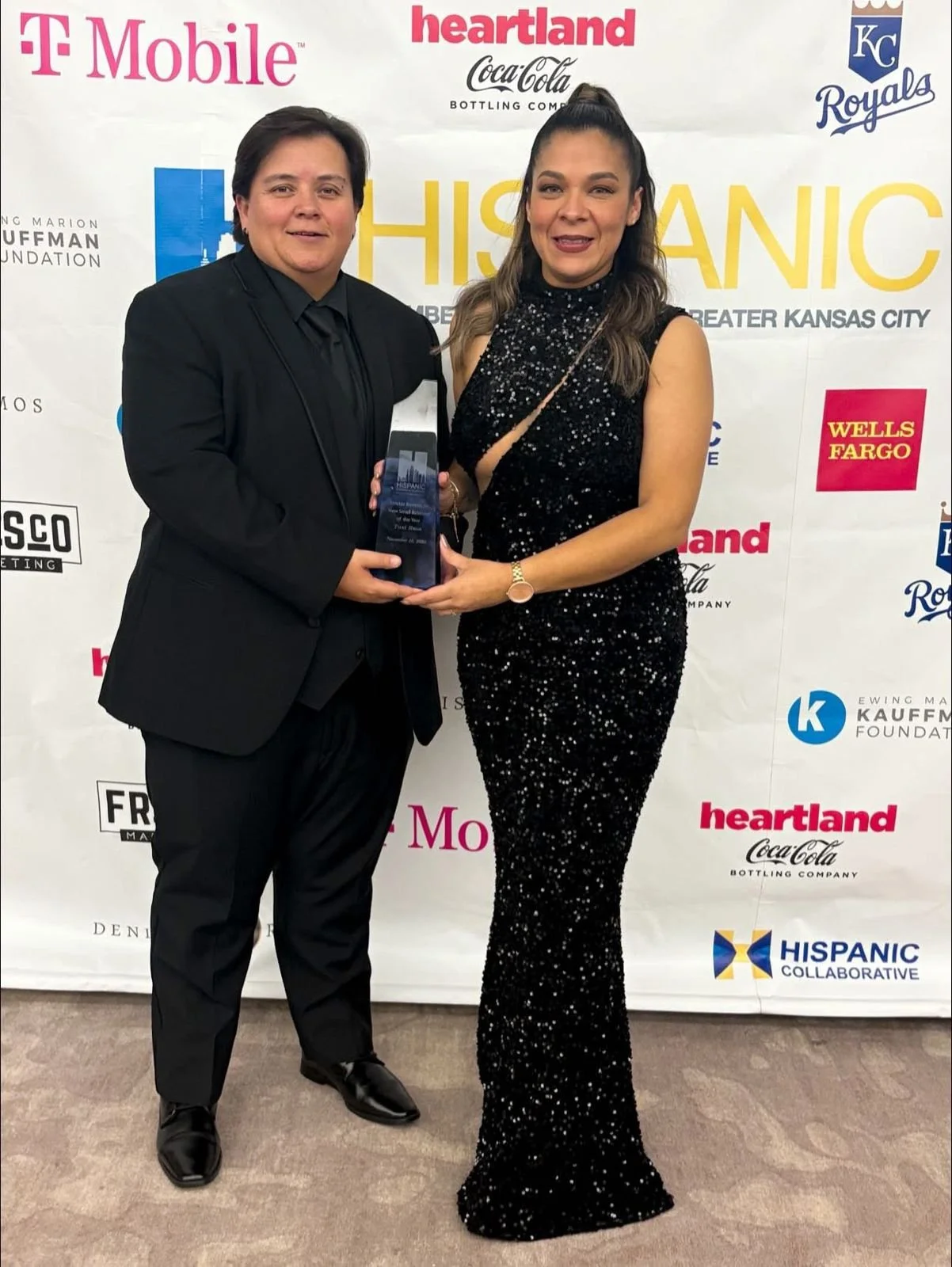 Dos personas vestidas de etiqueta en un evento, una mujer entregando un premio a un hombre, fondo con logotipos de marcas y organizaciones