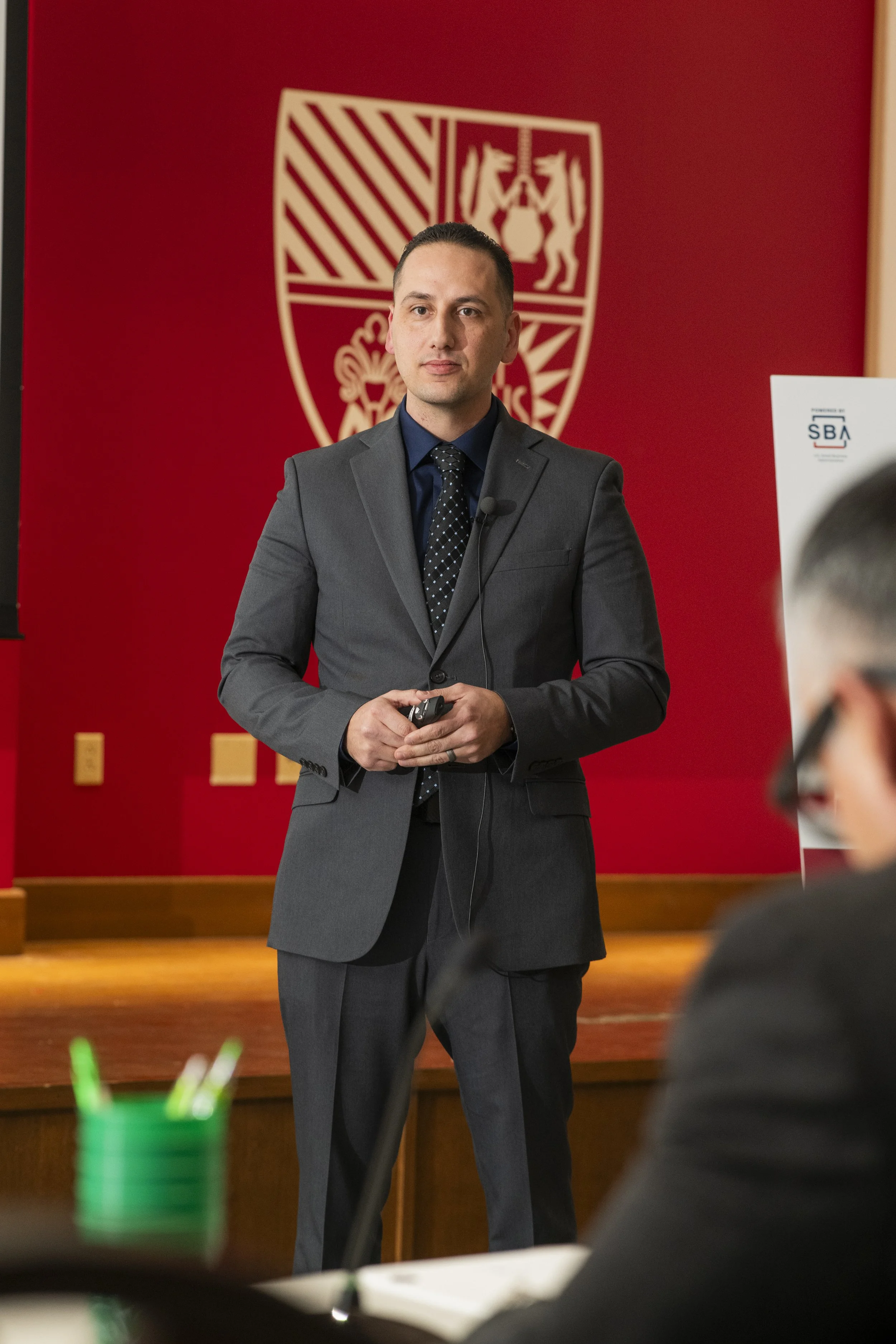 A man in a gray suit and tie stands in front of a red background with a white emblem. He has short dark hair and is holding a remote control, possibly giving a presentation or speech. The emblem behind him features two heraldic lions and a shield.