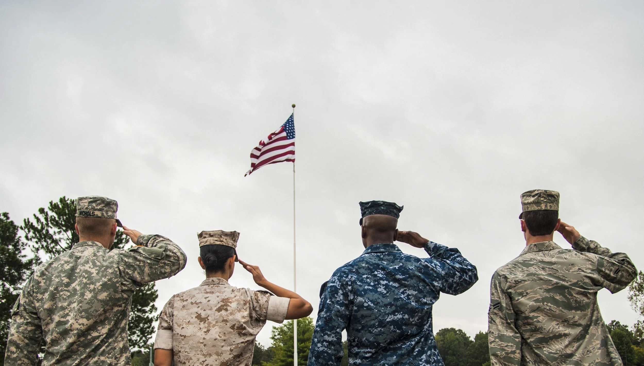 U.S. military personnel standing in formation, saluting, as the American flag is raised during a flag-raising ceremony outdoors under an overcast sky.