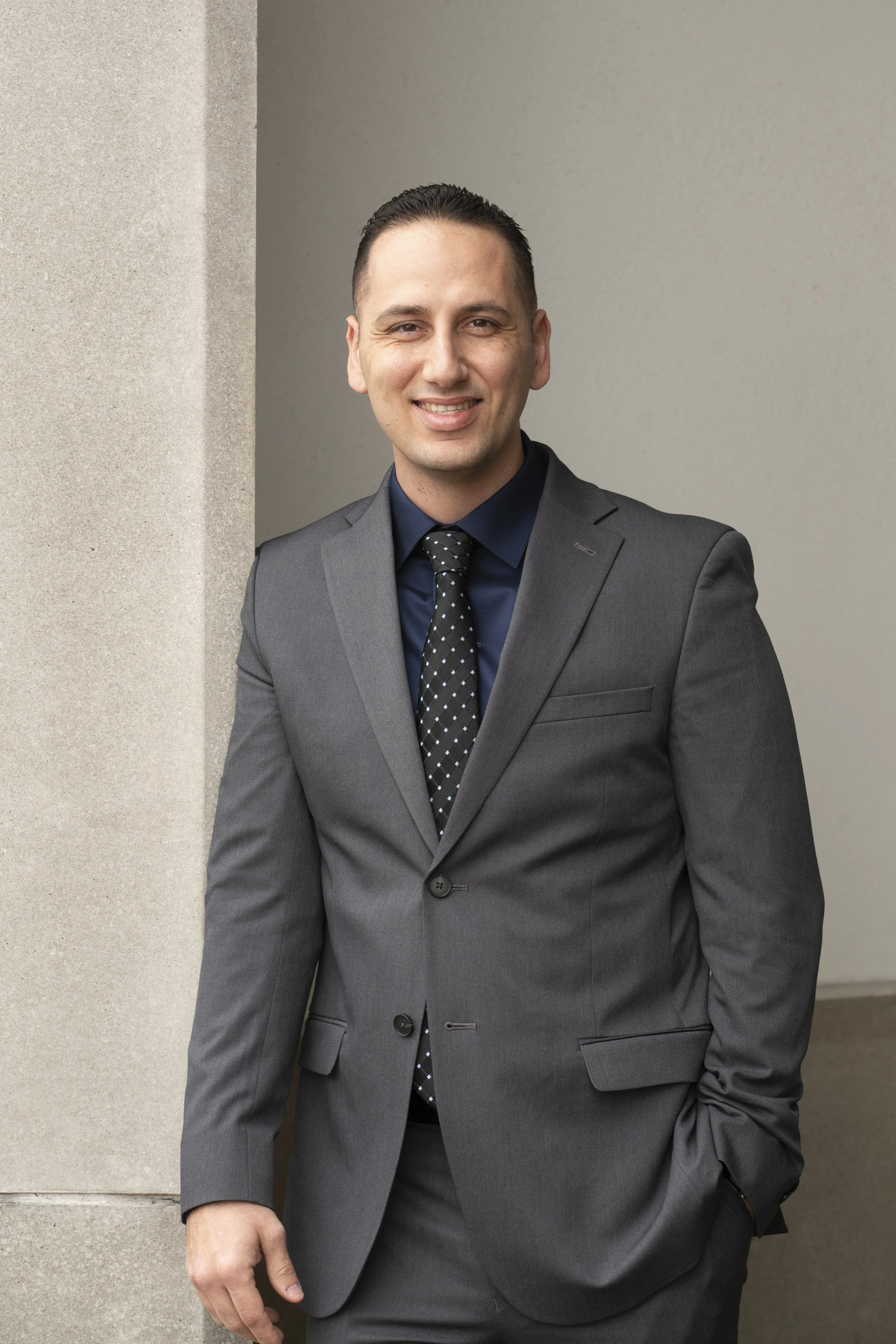 A man dressed in a dark gray suit with a black shirt and black dotted tie is standing outdoors against a beige wall. He is smiling and has short, dark hair, with one hand in his pocket and the other relaxed by his side.
