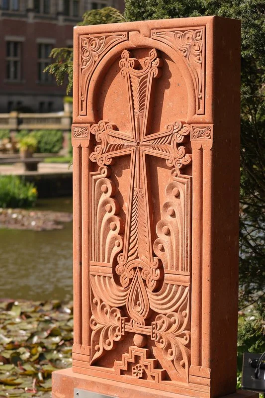 A carved stone or concrete religious cross with intricate designs, situated outdoors near water and greenery.