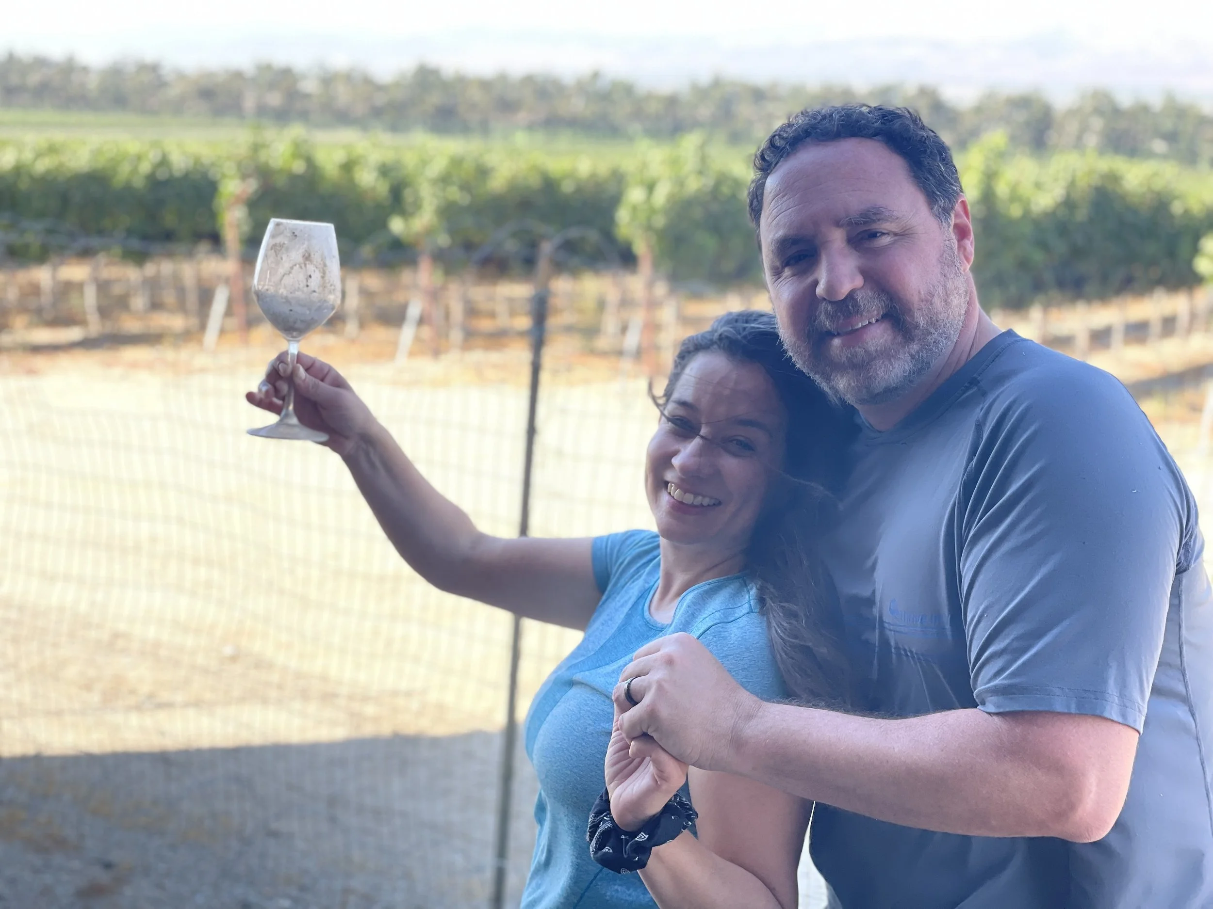 A smiling woman and man hugging outdoors, with the woman holding a wine glass, in front of a lush vineyard.