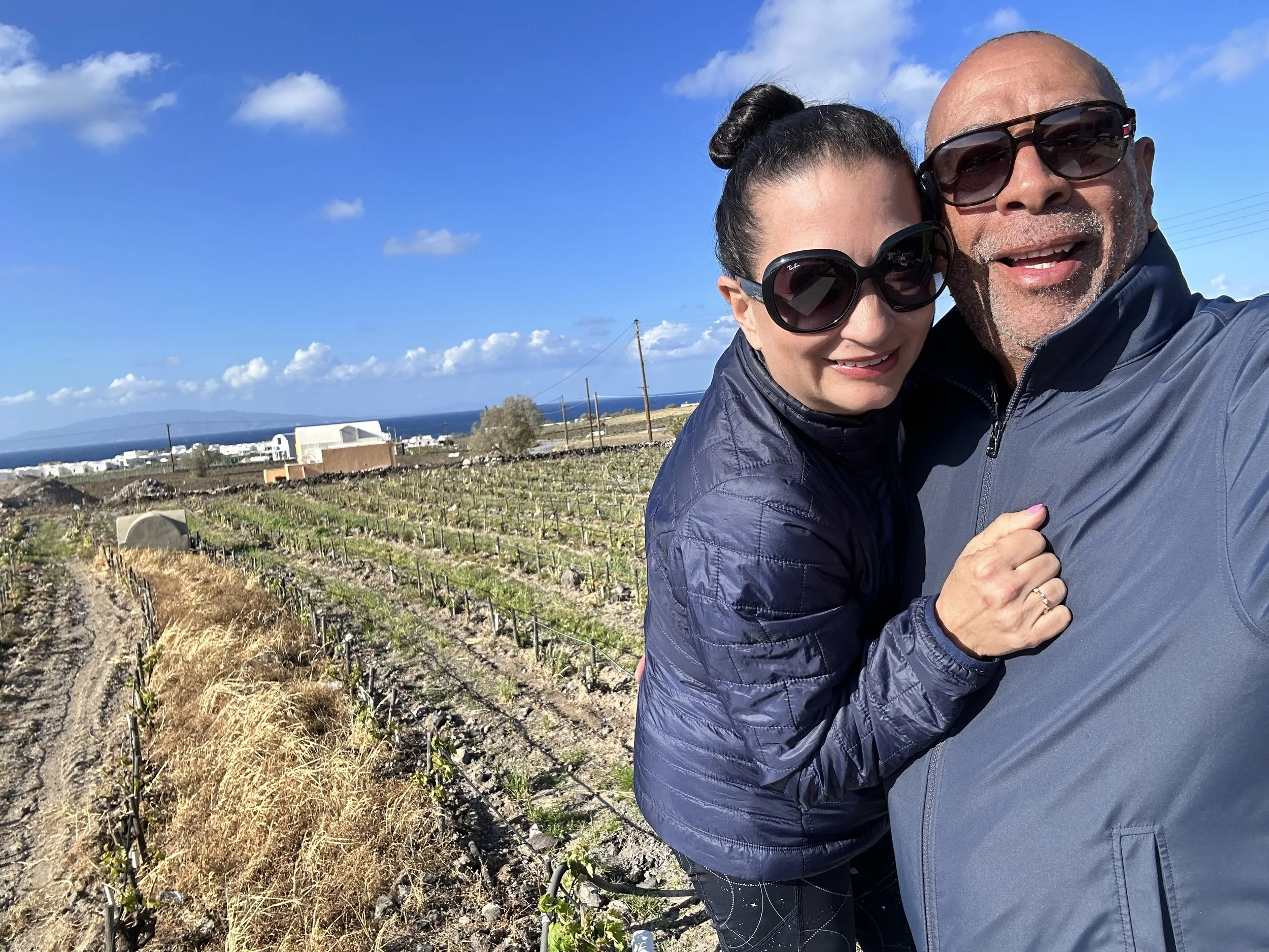 A couple taking a selfie outdoors, with a scenic view of a field, buildings, and the ocean in the background on a clear, sunny day.