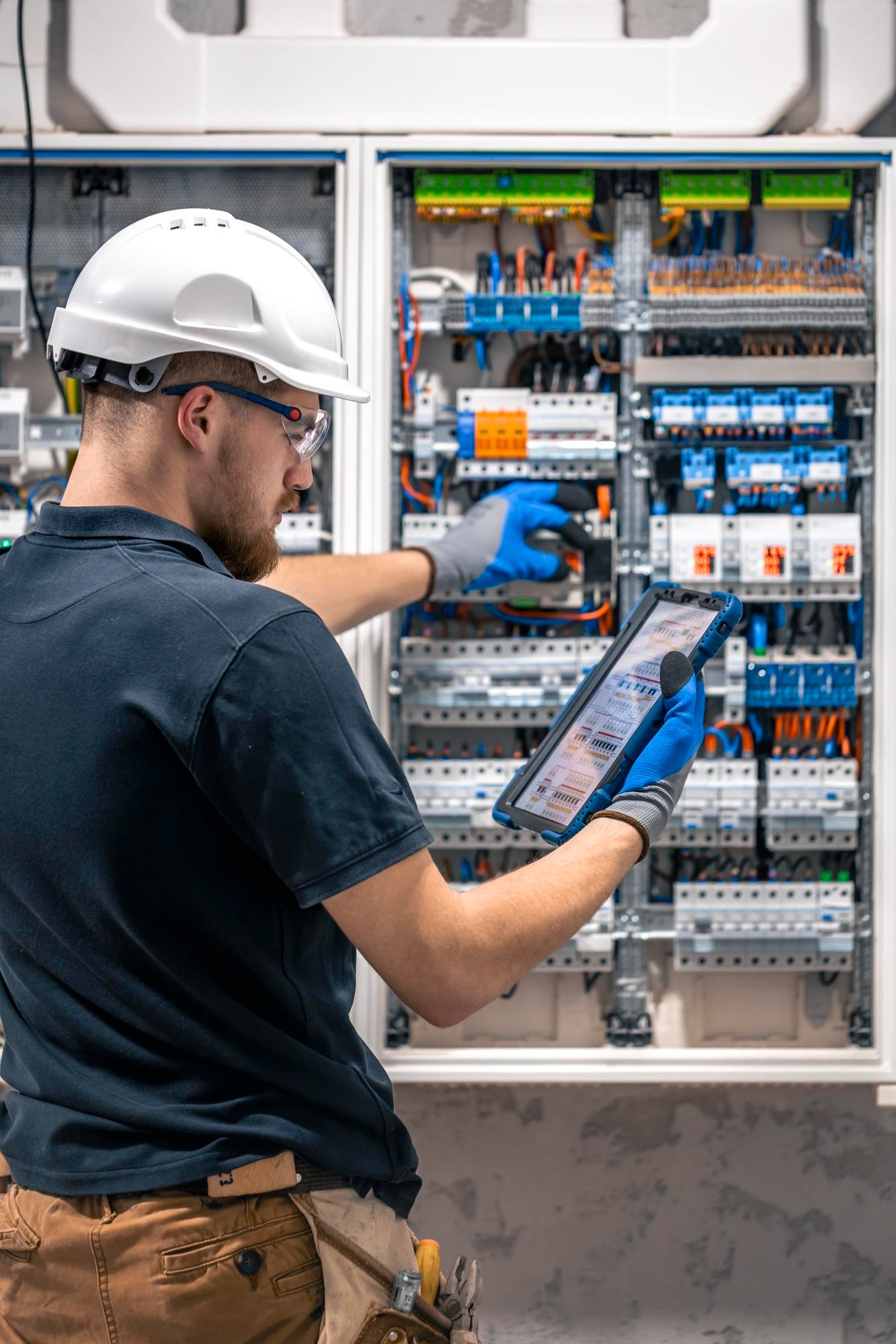 An electrician wearing a white safety helmet, safety glasses, gray gloves, and a dark shirt is working on an electrical panel with various wires and components. The electrician is holding a tablet device and appears to be troubleshooting or inspecting the electrical system.