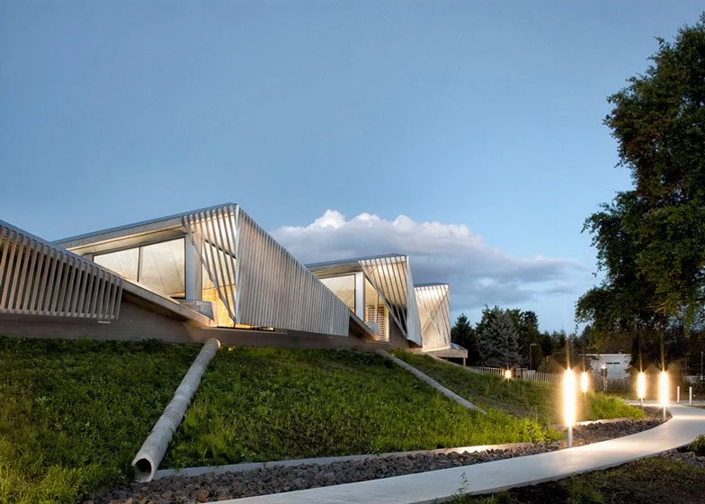 Modern building with angular architecture on a grassy hill, illuminated pathway with lights, trees, and a partly cloudy sky in the background.