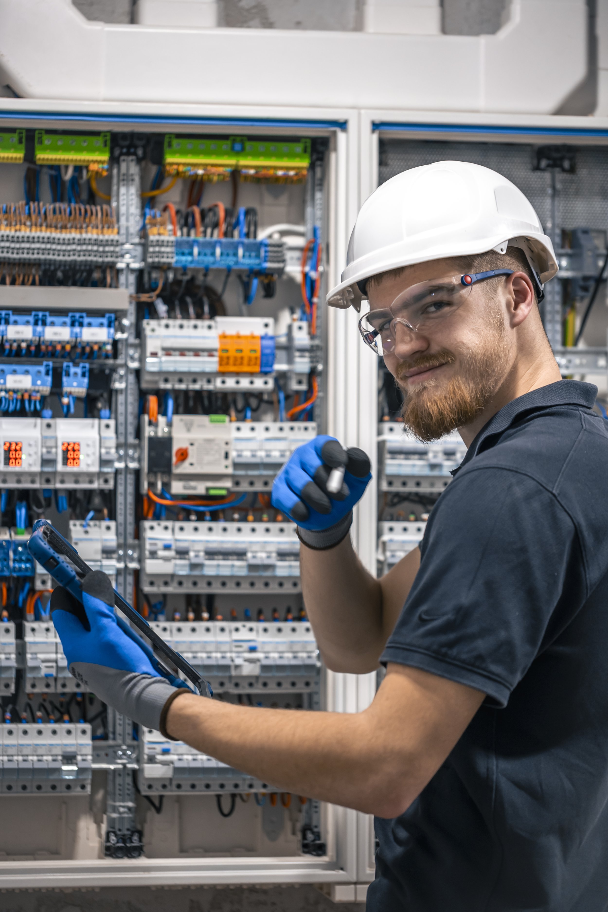 A male electrician wearing safety goggles, a white hard hat, and blue gloves working on an electrical control panel with many wires and circuit breakers.