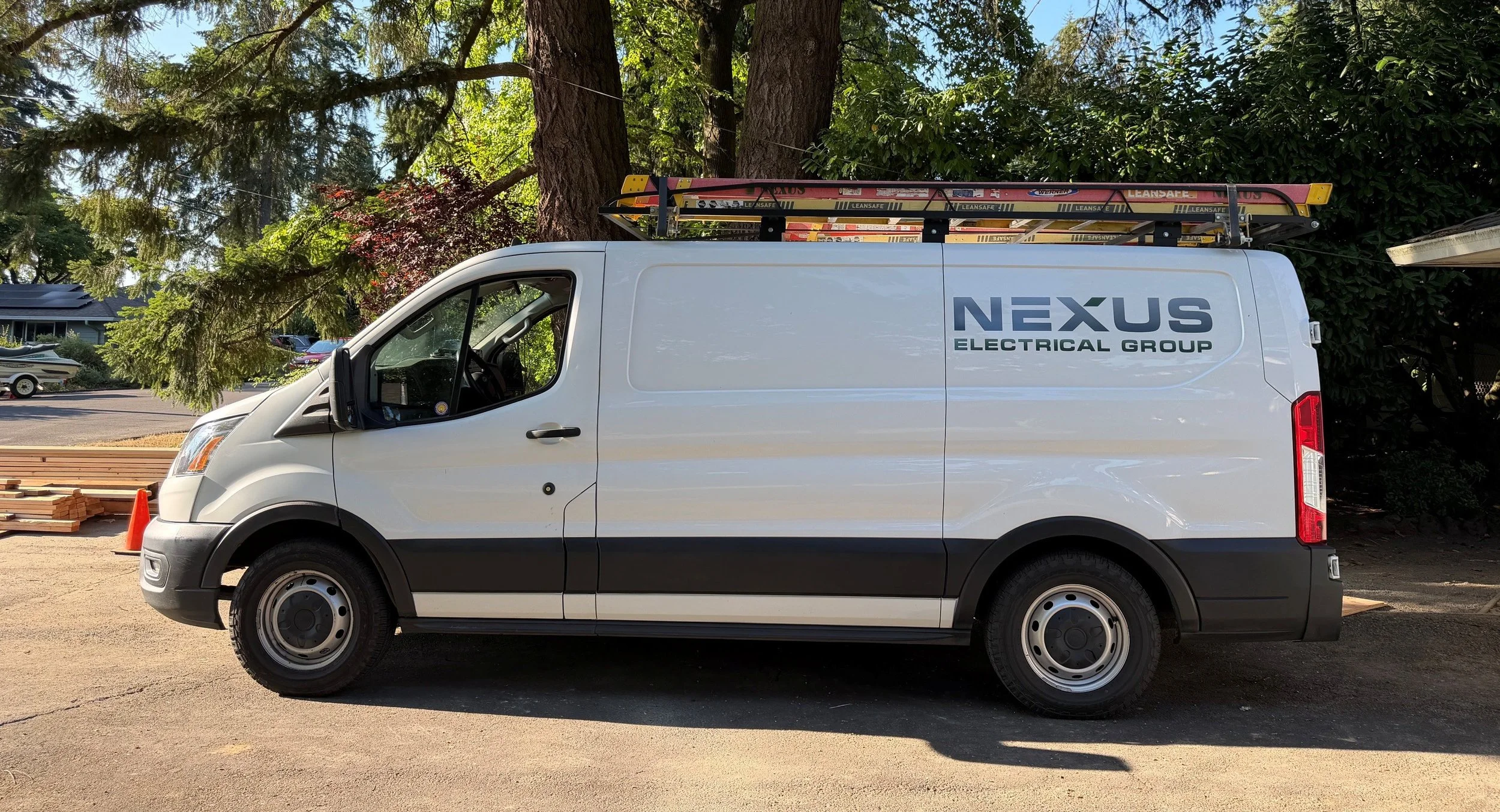 White Nexus Electrical Group service van parked outdoors near trees, with ladders mounted on top and orange safety cone nearby.