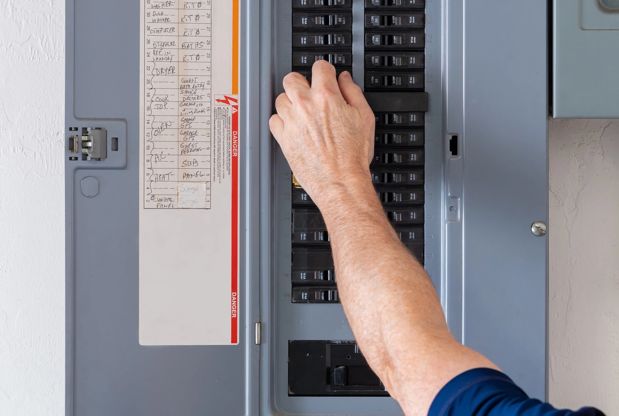 A person's hand is flipping a circuit breaker switch in an electrical panel, which is mounted on a white wall.