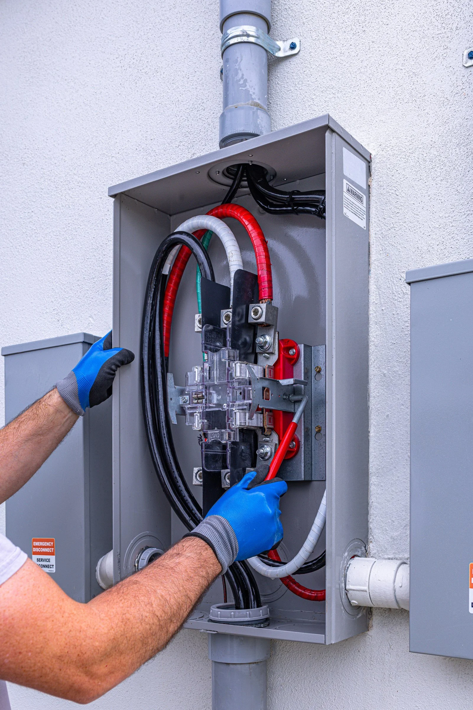 A technician wearing blue gloves working on an electrical panel outside a building, with wiring and components visible.