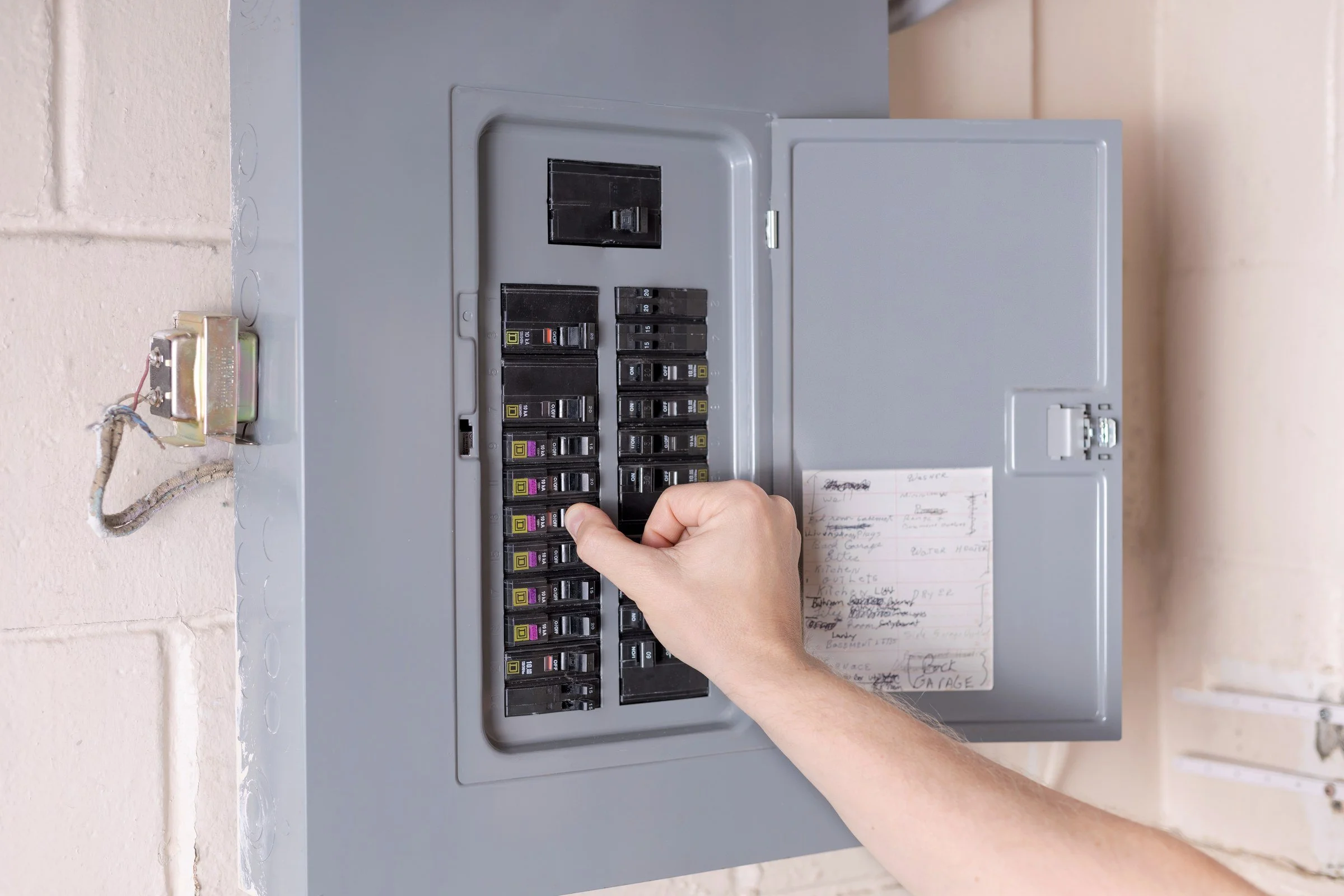 A person with light skin and no visible clothing on the arms operates a gray electrical circuit breaker panel with multiple switches, some of which have labels and colors, attached to a wall.