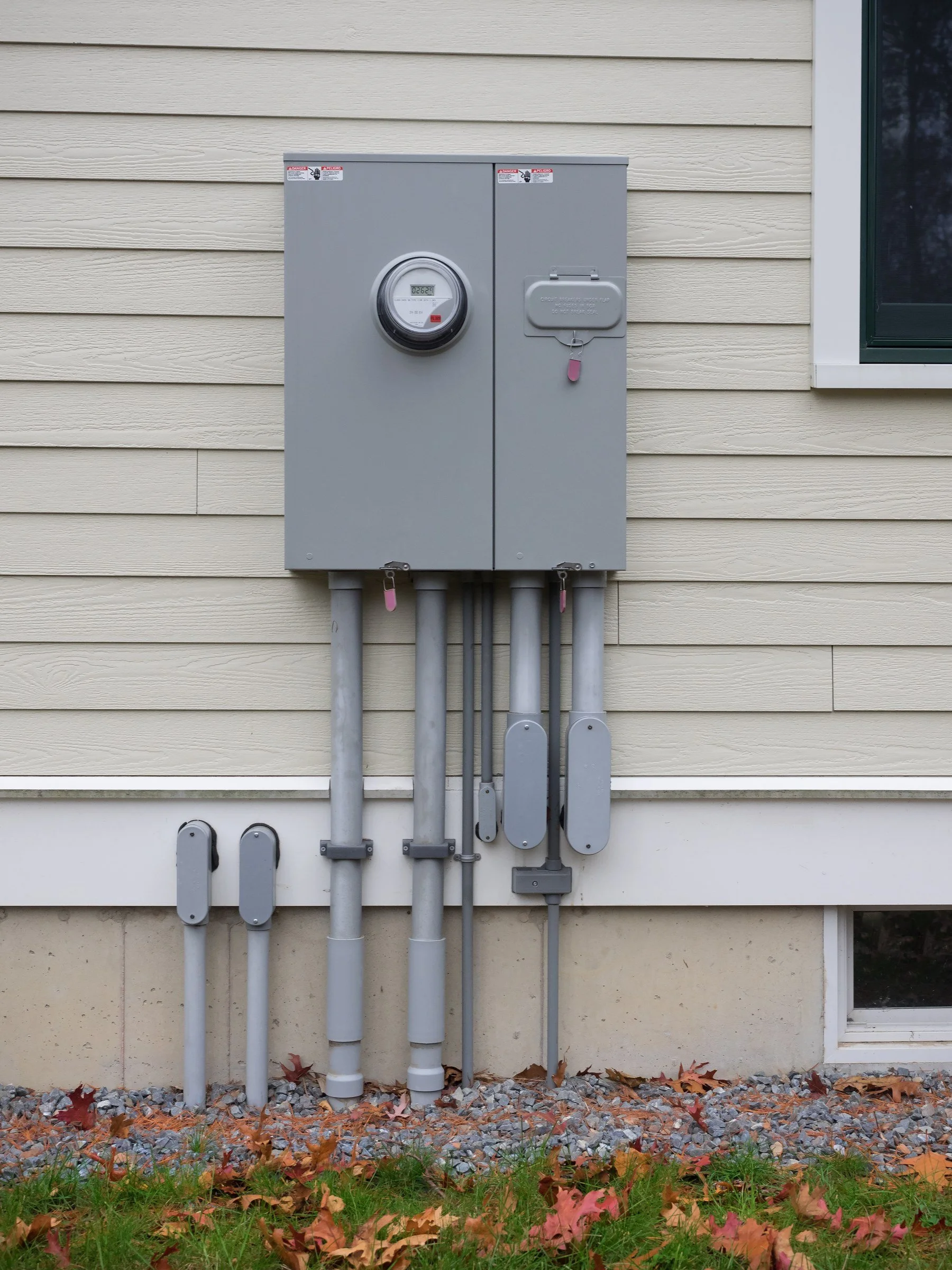 Exterior view of an electric utility box mounted on a house wall, with several electrical conduits extending downward and autumn leaves scattered at the base.