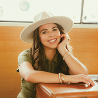 A woman in a green outfit and white hat smiles warmly, sitting next to two stacked vintage brown suitcases in a cozy Volkswagen van interior with wood paneling.