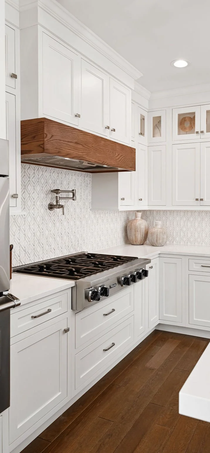 White kitchen with upper and lower cabinets, a gas stove, a wooden range hood, decorative vases on the counter, and a textured backsplash.