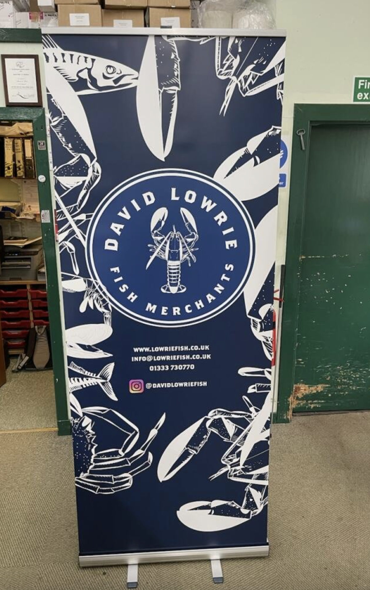 Promotional display for David Lowrie Fish Merchants with a circular logo featuring a lobster and the text 'David Lowrie Fish Merchants'. Contact information and social media handle are also visible.
