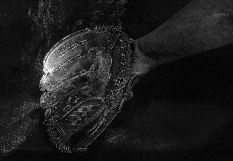 A person wearing a baseball glove in a focused catch or throw position, with a dark background.